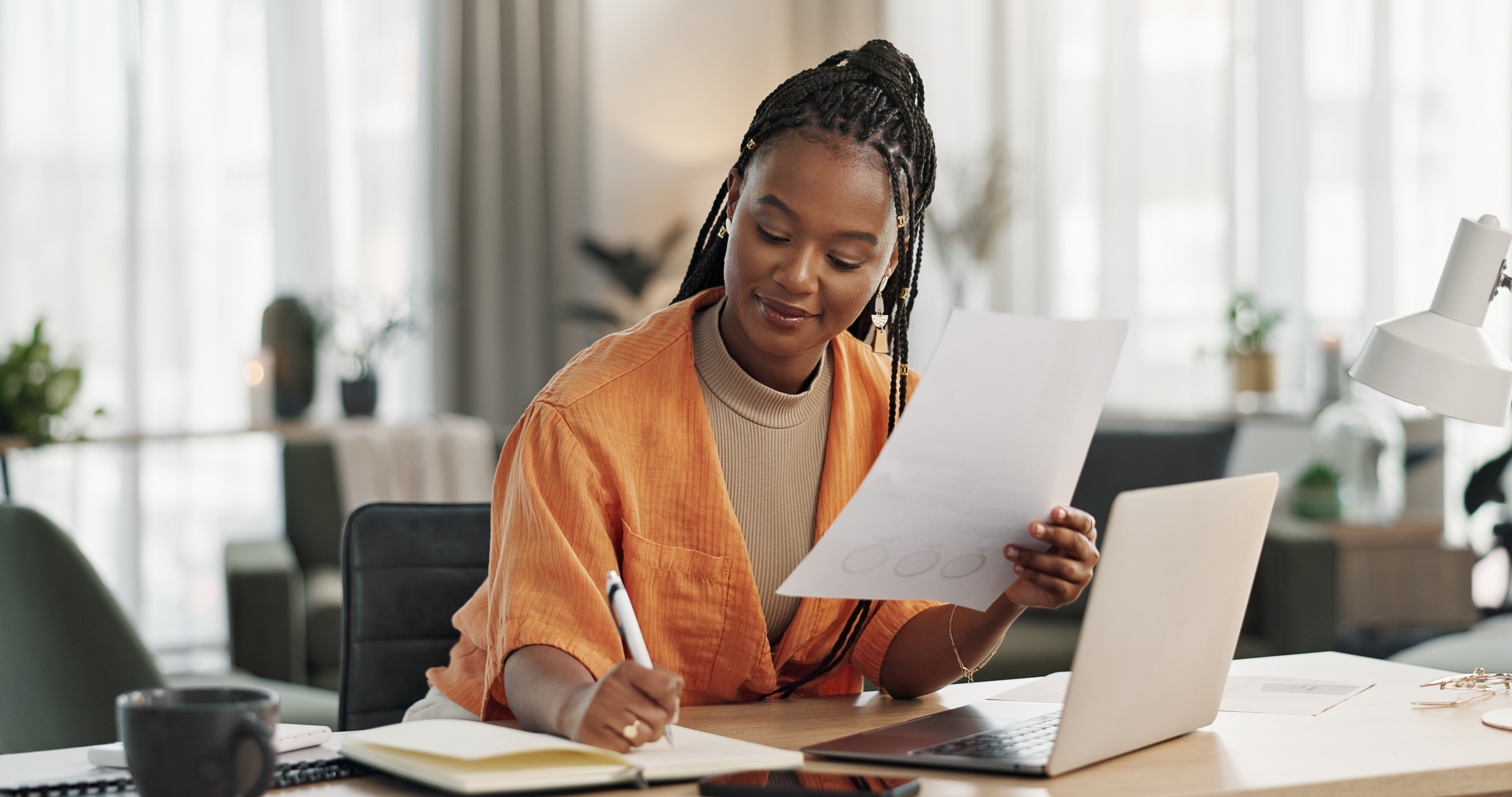 Woman in orange shirt writing notes while holding paper, working at desk with laptop and coffee mug.