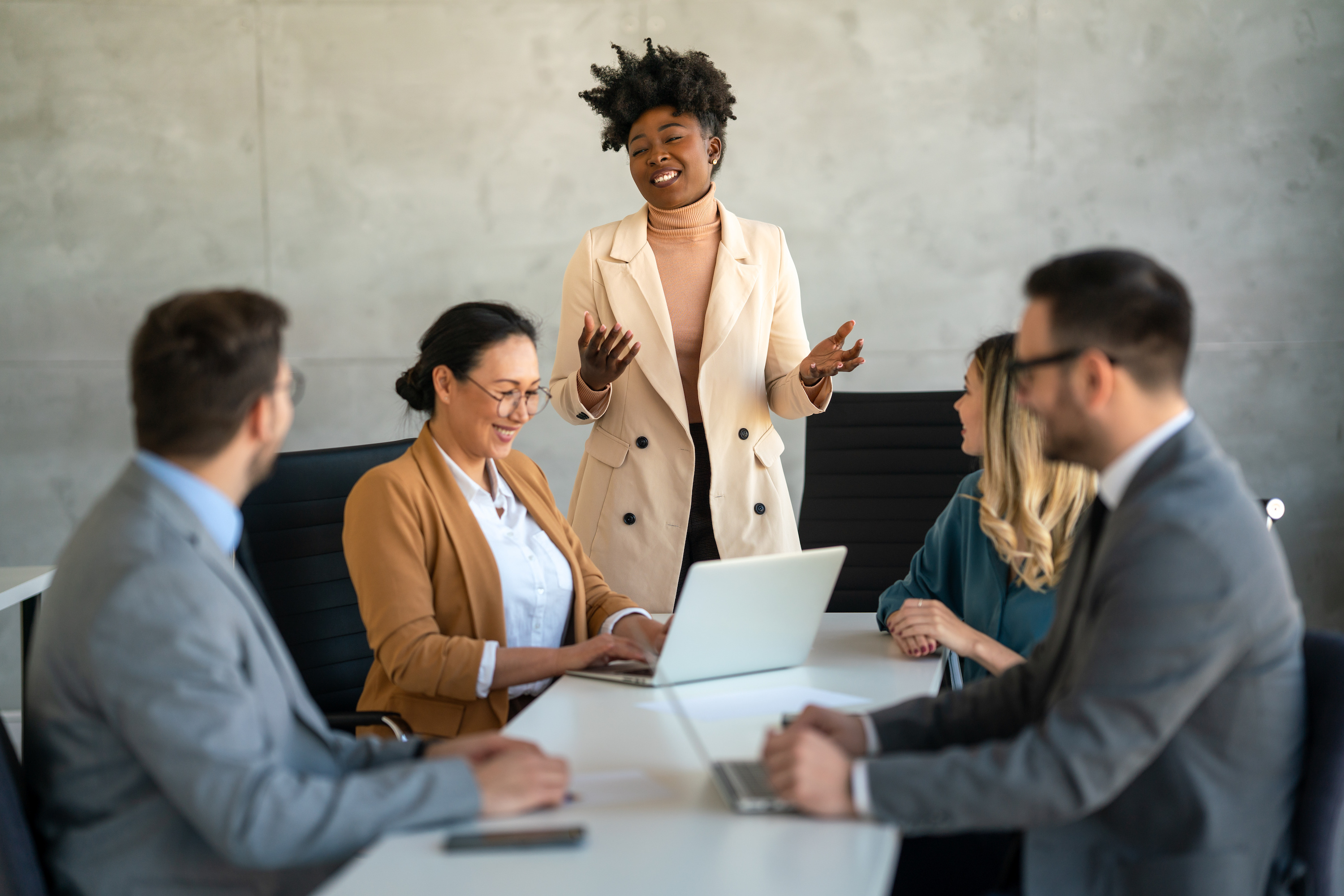 Businesswoman standing and speaking to three seated coworkers around a conference table with laptops.