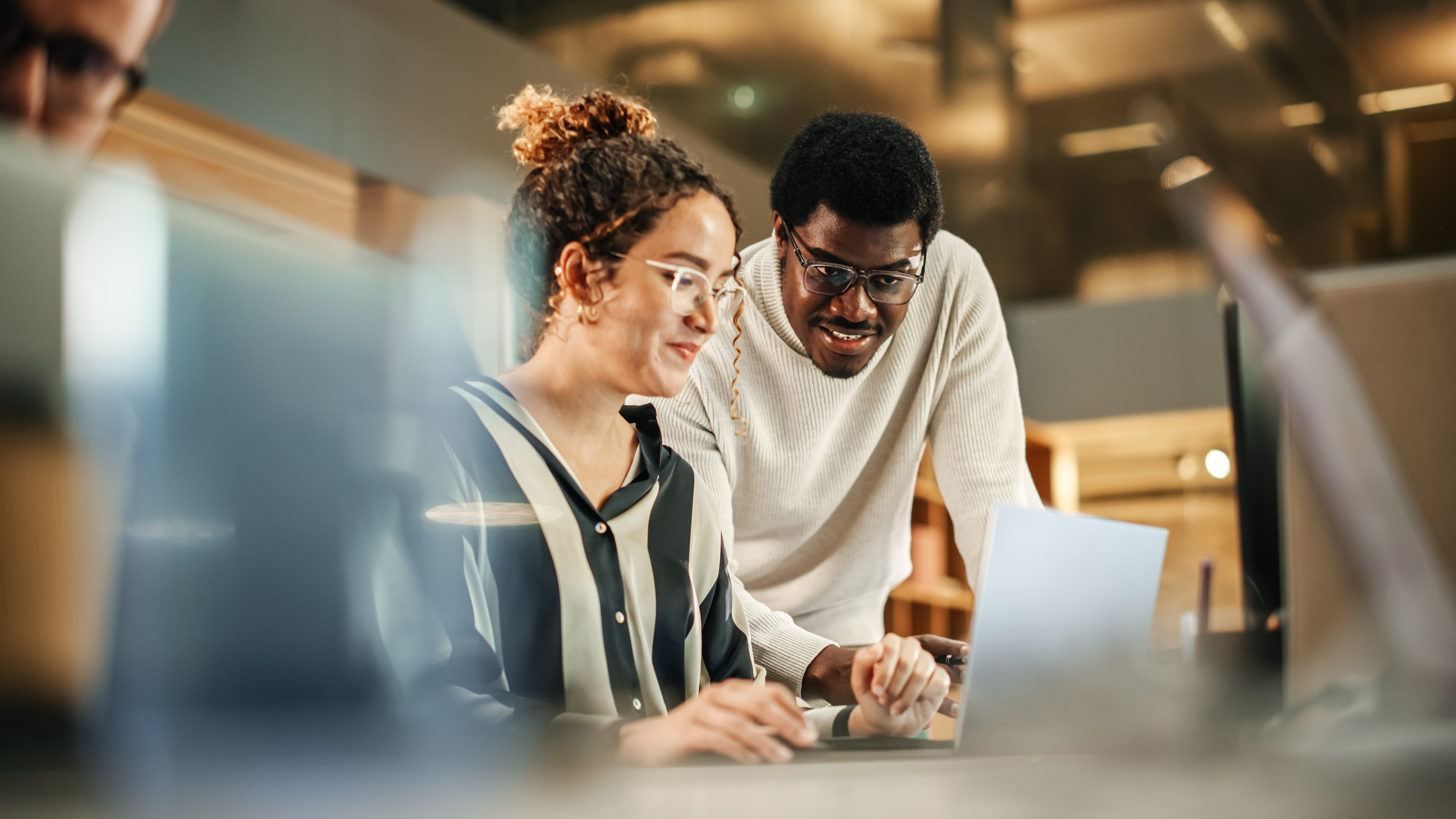 Man in glasses leans over woman typing on laptop in an office setting.