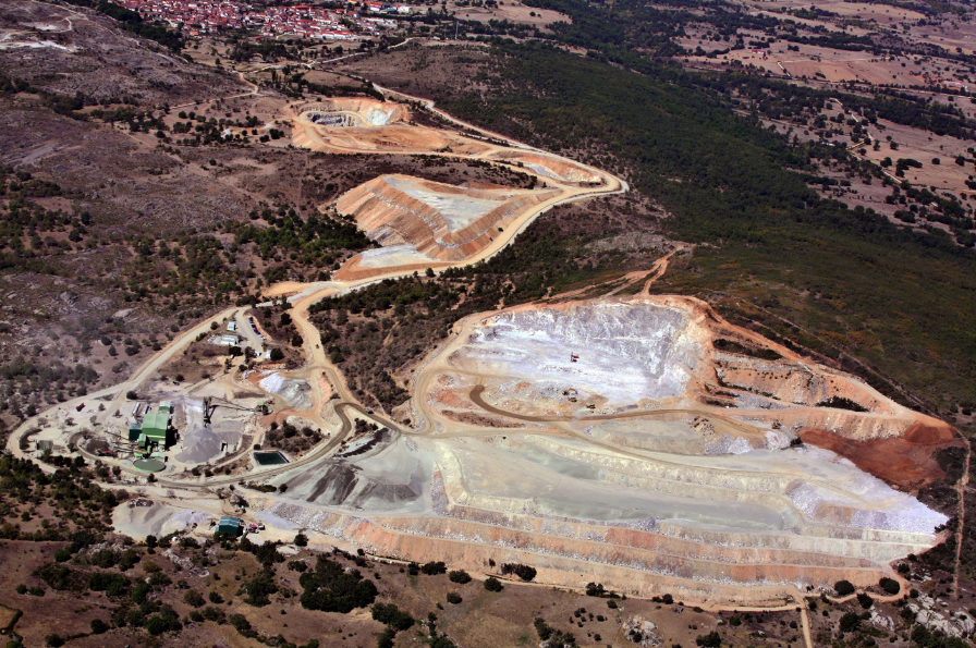 Aerial view of a large open-pit tungsten mining site with terraced excavation, surrounding forest, processing facilities, and nearby rural village.
