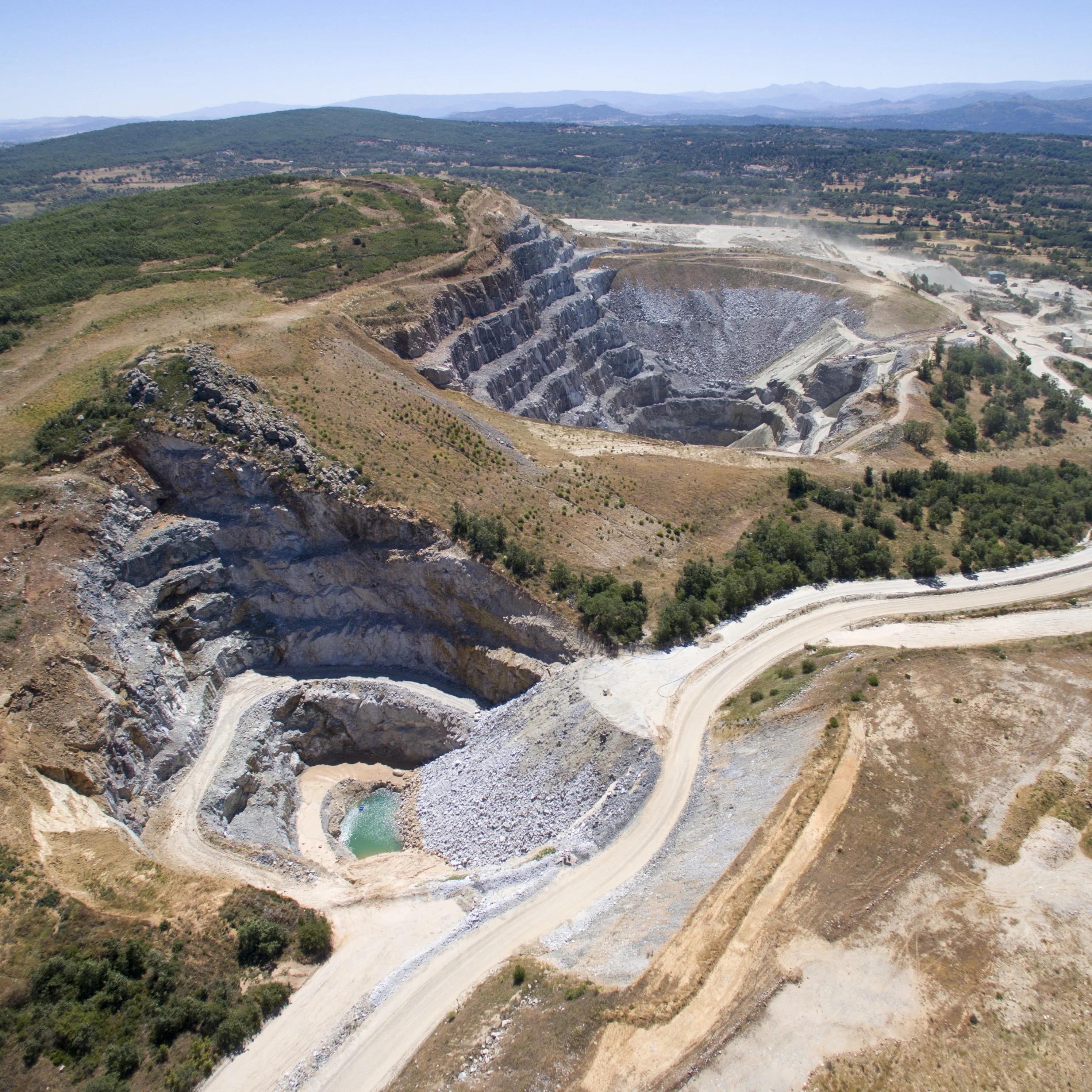 Aerial view of an open-pit mining operation surrounded by rugged terrain and sparse vegetation, with terraced rock layers and access roads.