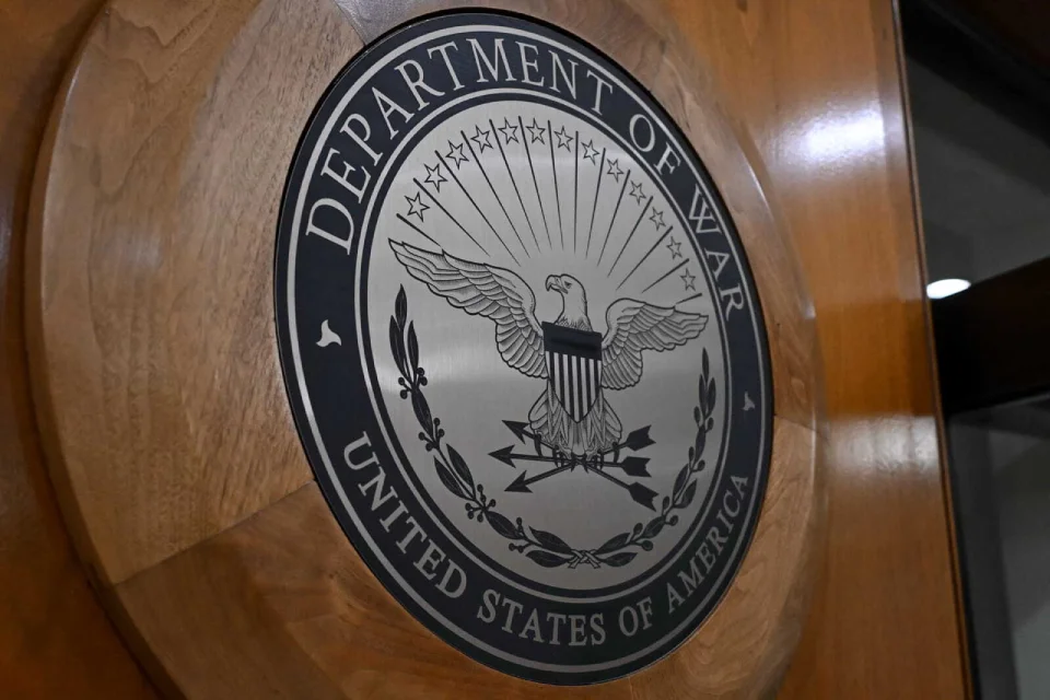 Seal of the United States Department of Defense displayed on a podium, with the Pentagon emblem and American flags in the background.
