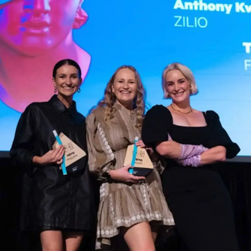 Three women smiling and posing on stage at an awards event, two holding trophies.