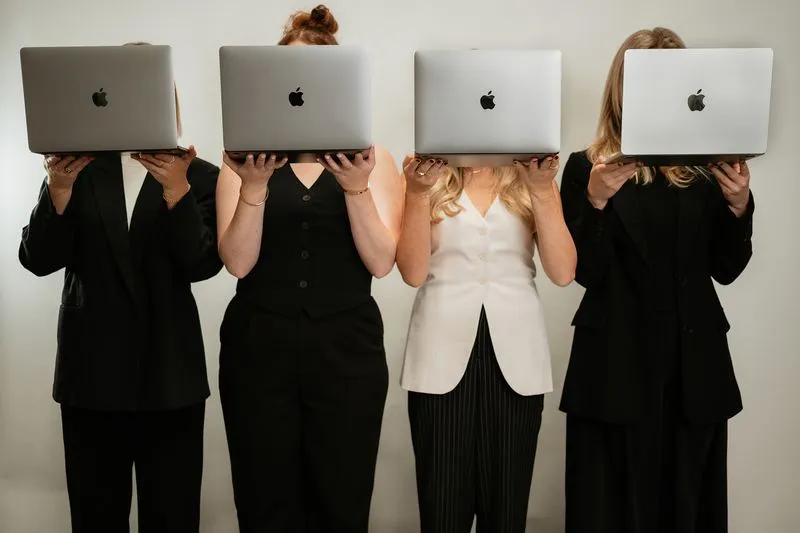 Four businesswomen standing side by side holding MacBook laptops in front of their faces.
