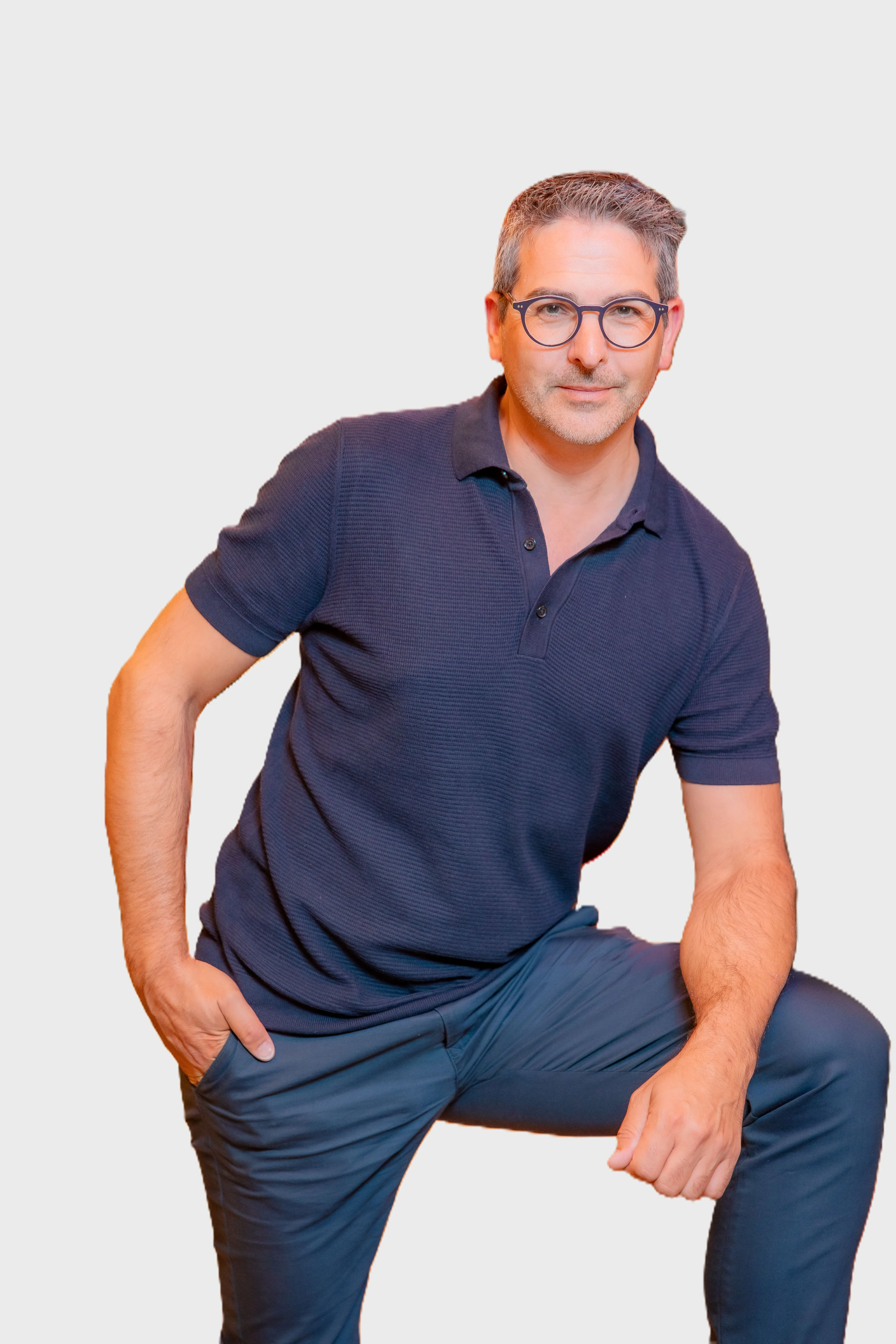 Mann mittleren Alters mit Brille und grauem Haar trägt ein dunkelblaues Poloshirt und blaue Hose, eine Hand in der Hosentasche, kniet leicht.