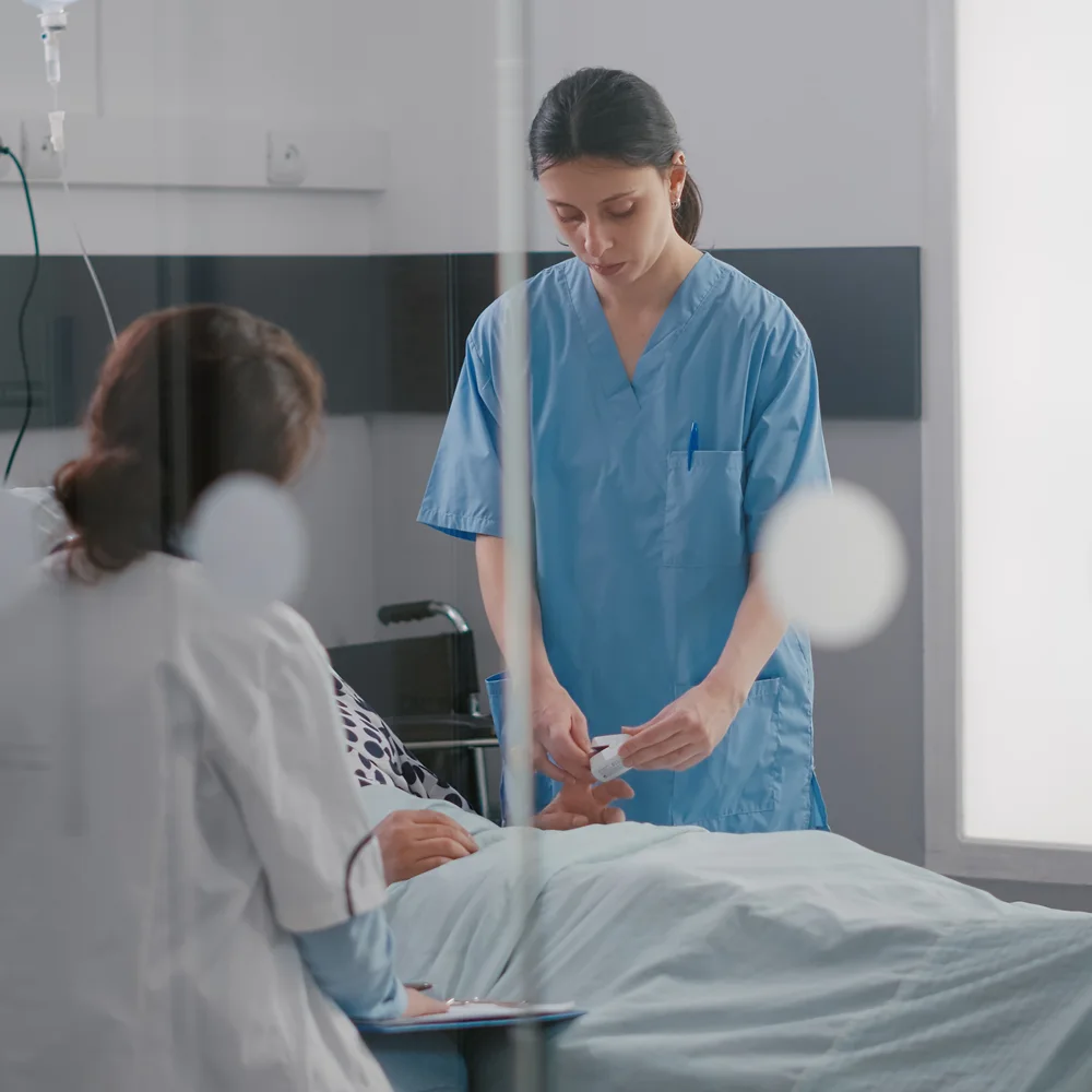 Nurse in blue scrubs administers care to a hospital patient while a doctor takes notes nearby, reflecting attentive inpatient care and teamwork in healthcare settings.