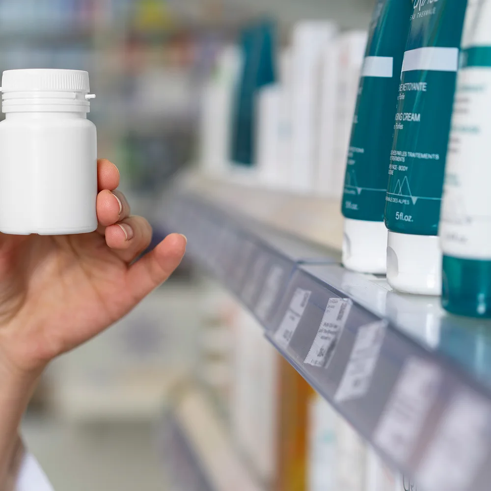 Close-up of a person selecting a white plastic pill bottle from a pharmacy shelf, illustrating pharmaceutical services and over-the-counter medication access.