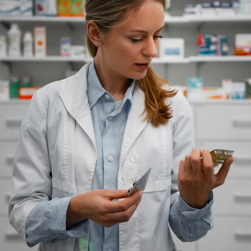 Female pharmacist examines different medication blister packs behind the pharmacy counter, representing professional prescription services and medication accuracy.
