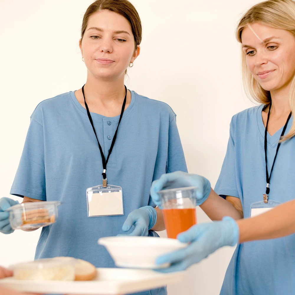 Two female caregivers in light blue uniforms distribute food and drinks to a patient, highlighting hospital meal service and patient support roles.