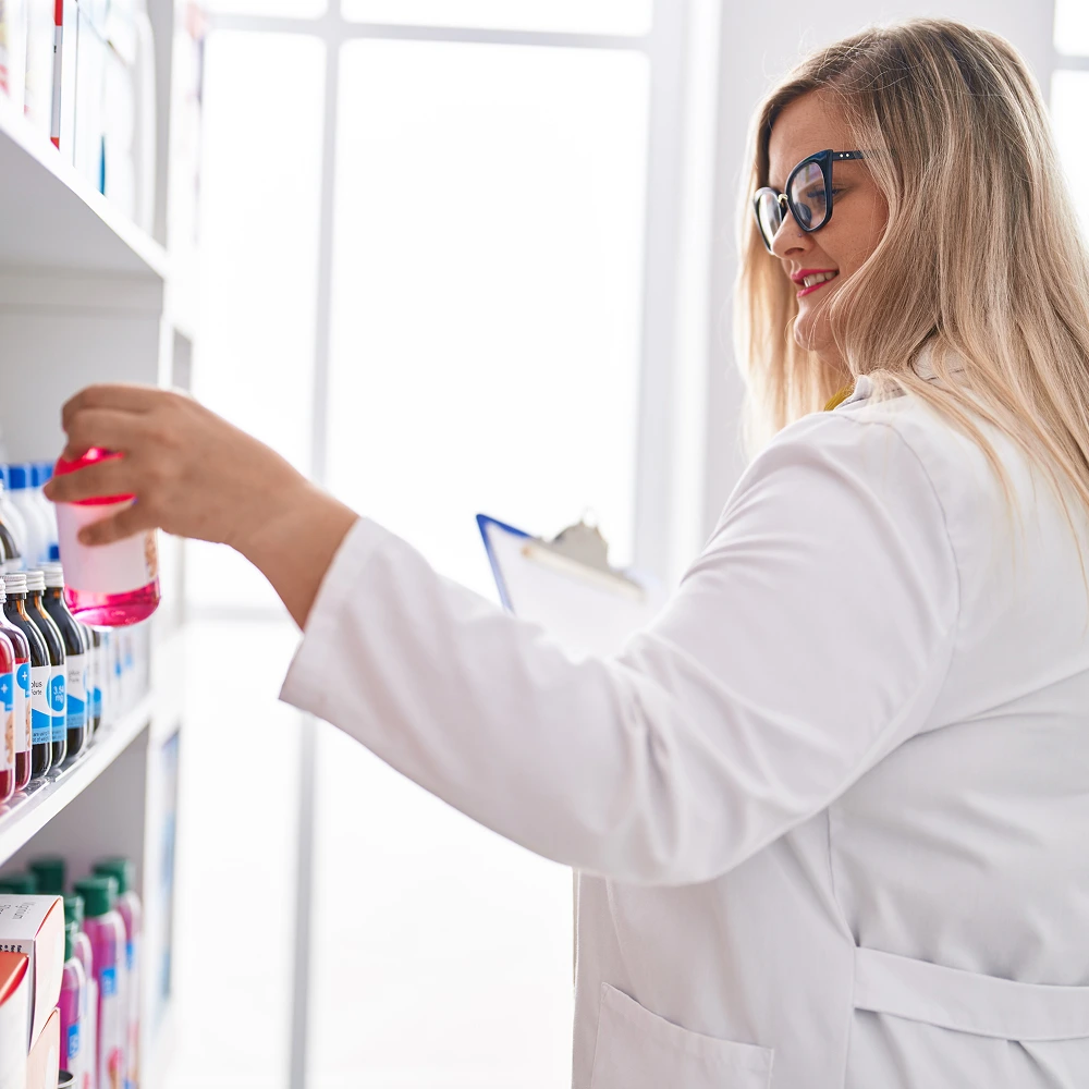 Smiling female pharmacist in a white coat checks products on a pharmacy shelf, holding a clipboard, symbolizing inventory management and pharmaceutical care.