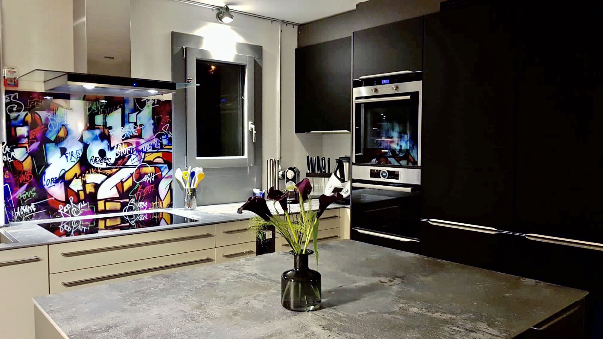 Modern kitchen with beige lower cabinets, black tall cabinets, built-in oven, and colorful graffiti backsplash.