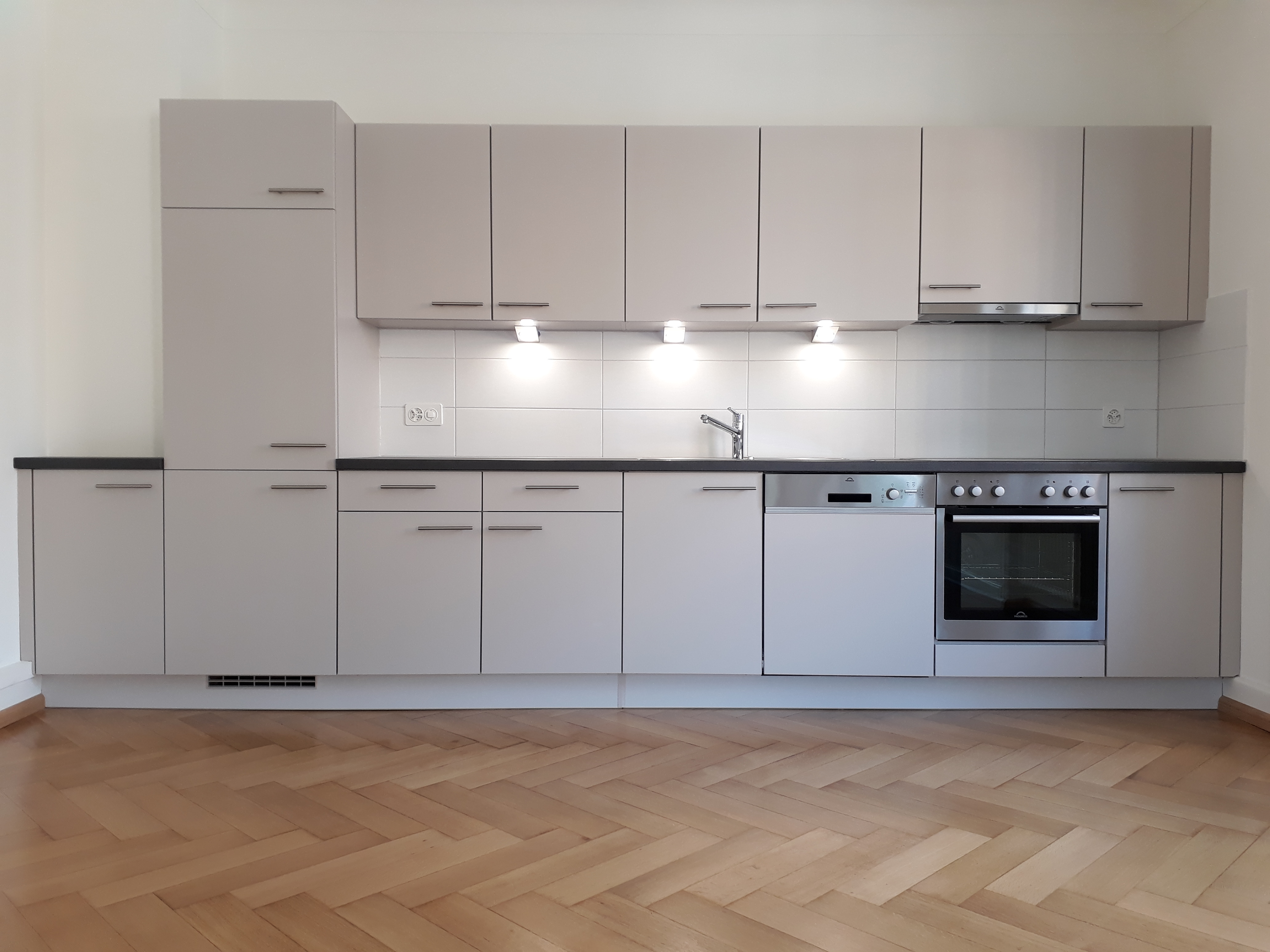 Modern minimalist kitchen with light gray cabinets, black countertop, built-in dishwasher, oven, stovetop, and under-cabinet lights.