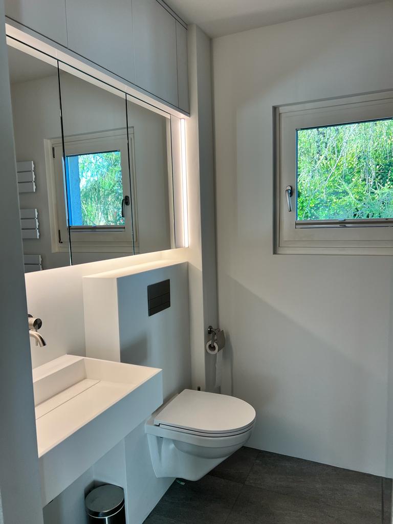 Minimalist white bathroom with a wall-mounted toilet, rectangular sink, mirror cabinet with lighting, and a window showing greenery outside.