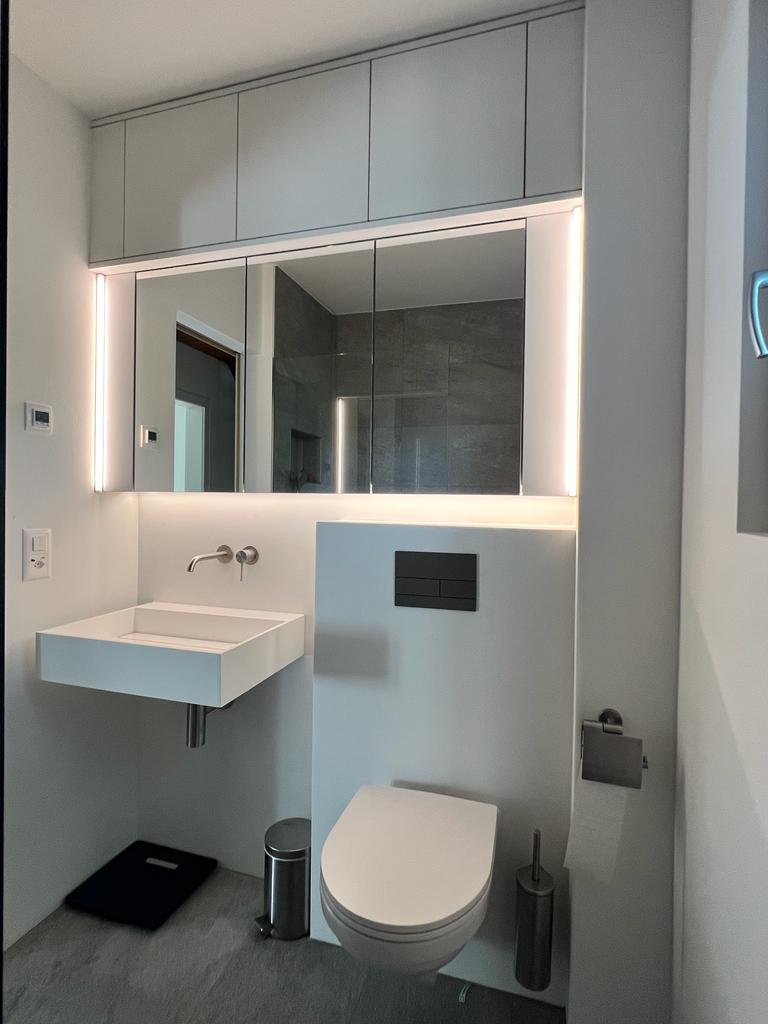 Minimalist bathroom with floating white sink, wall-mounted toilet, illuminated mirror cabinets, and gray tile floor.