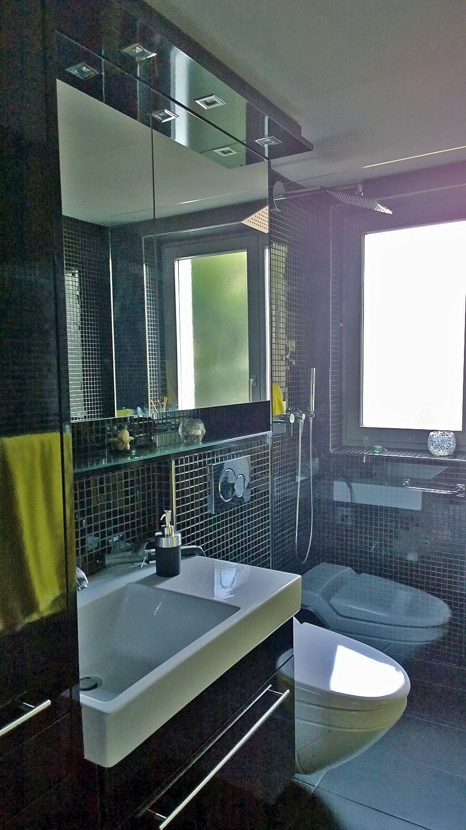 Modern bathroom with dark mosaic tiles, white rectangular sink, wall-mounted toilet, and large frosted window.