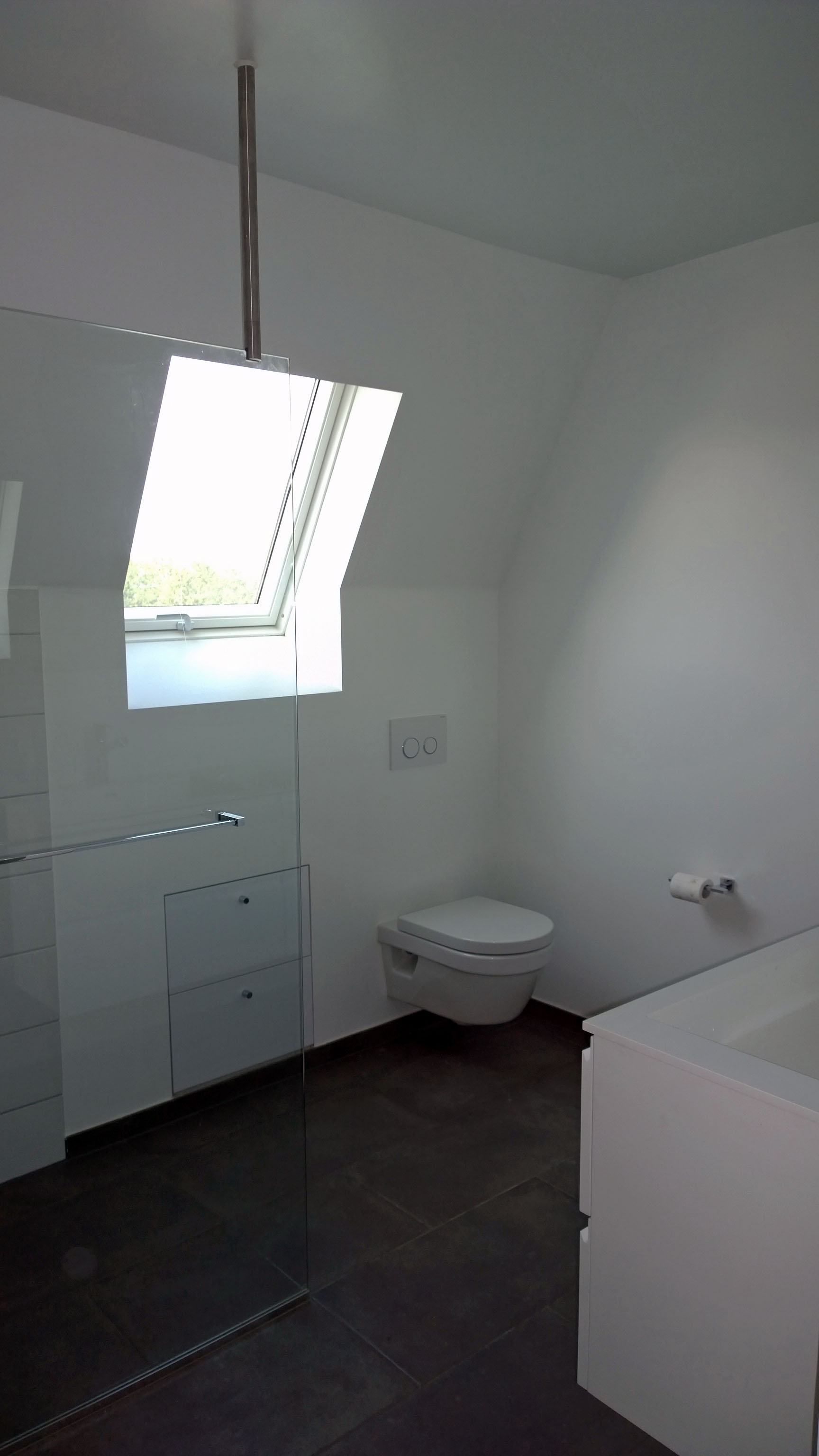 Minimalist bathroom with a wall-mounted toilet, a bathtub, dark floor tiles, and a window on a slanted ceiling.