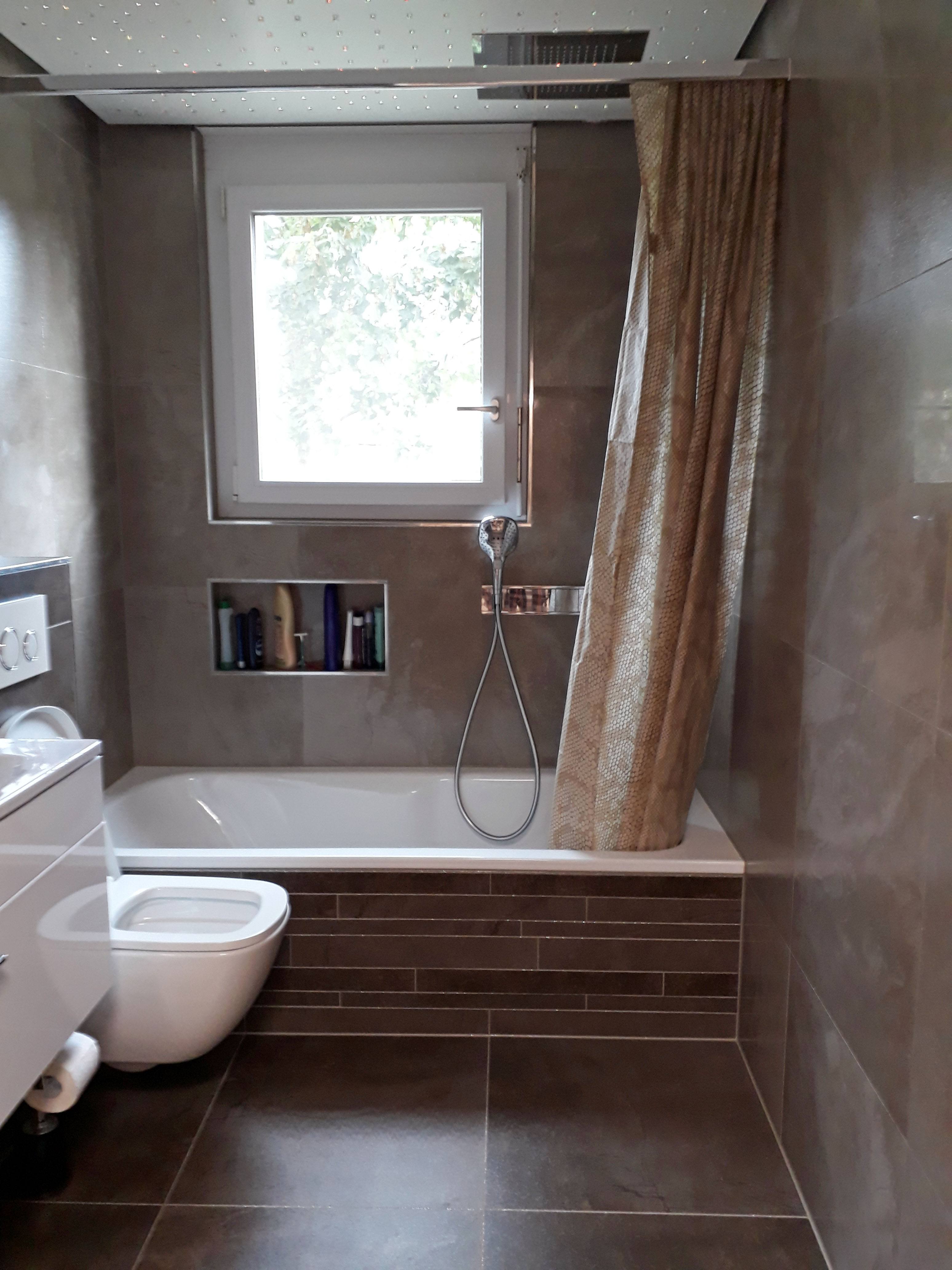 Modern bathroom with a white bathtub, showerhead, beige shower curtain, wall-mounted white toilet, and a window above the tub.