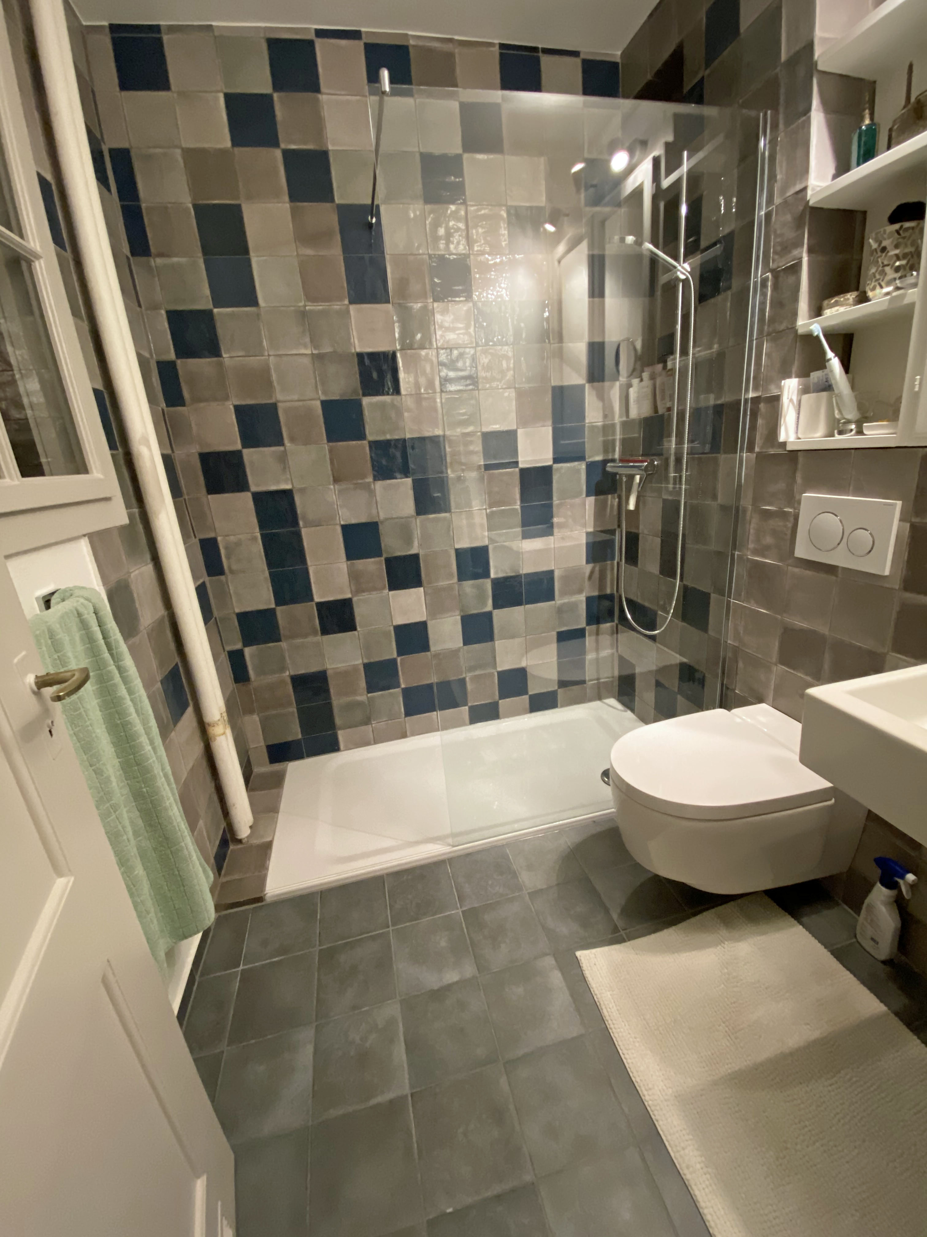 Modern small bathroom with a glass shower enclosure, blue and gray tiled walls, a wall-mounted toilet, and a white sink with shelves.