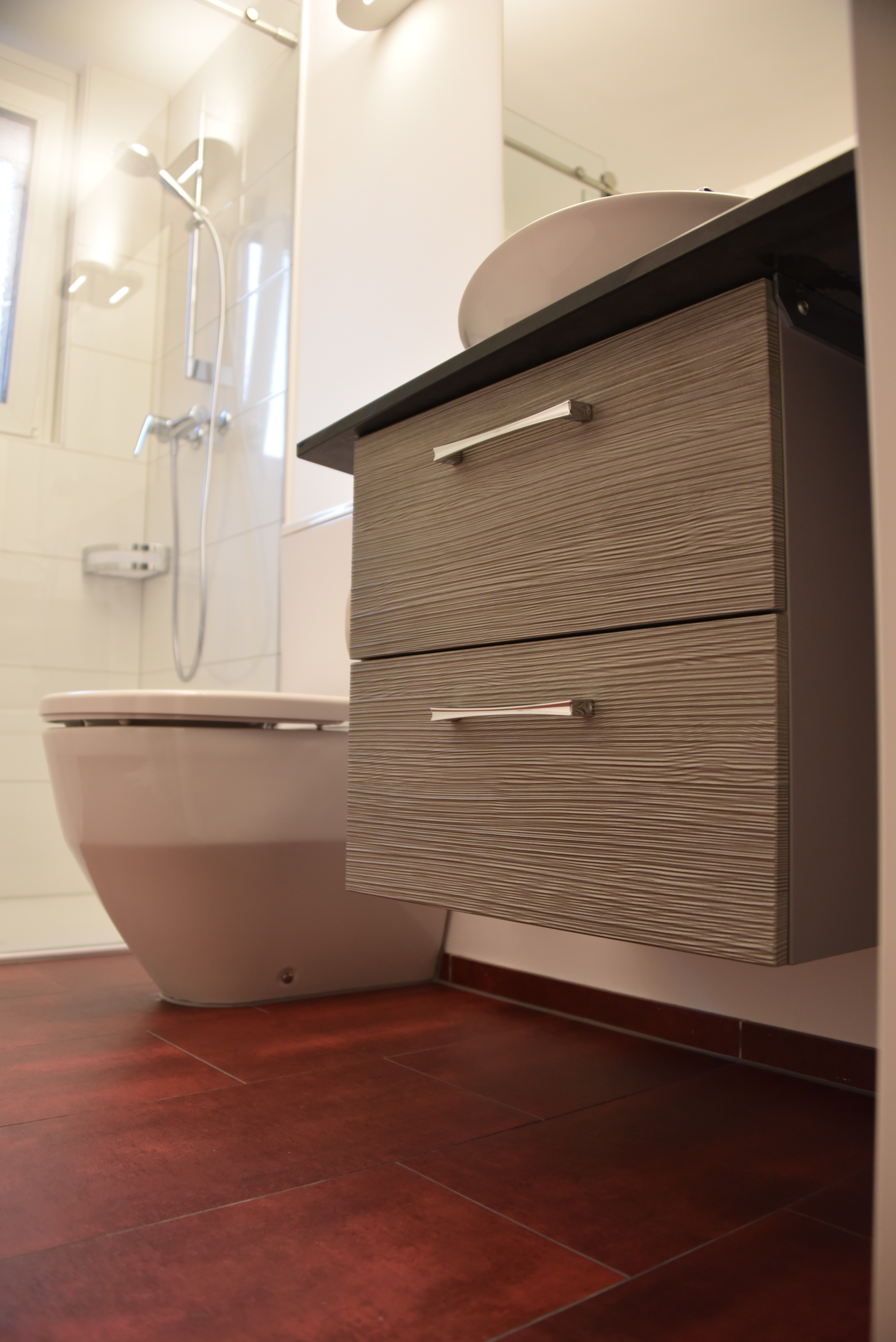 Modern bathroom interior with a wall-mounted wood-textured vanity, round sink, toilet, and glass shower enclosure with a showerhead.