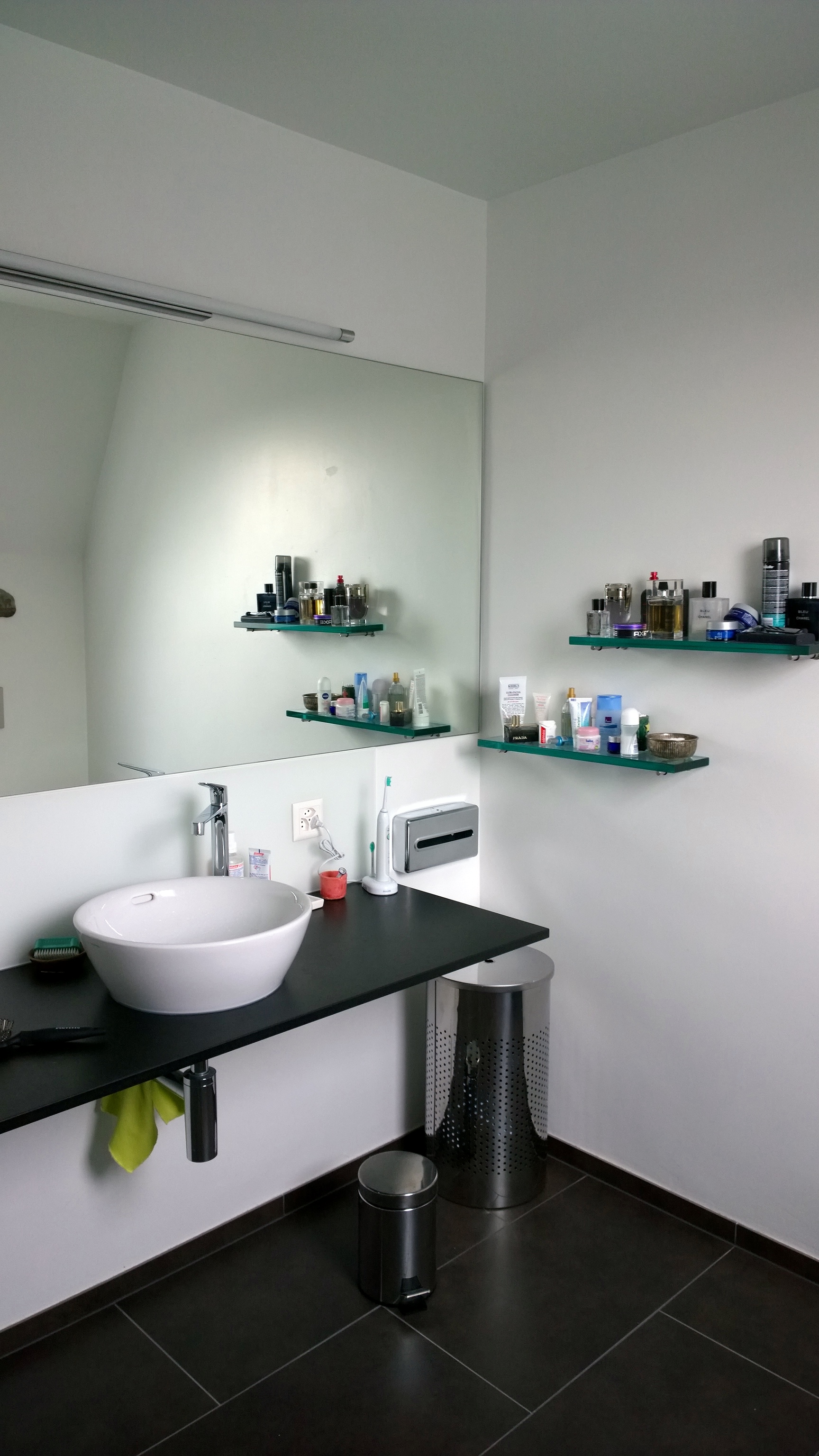 Modern bathroom sink with a round white basin on a black countertop, small shelves with toiletries on the wall, and two stainless steel trash bins on dark tiled floor.