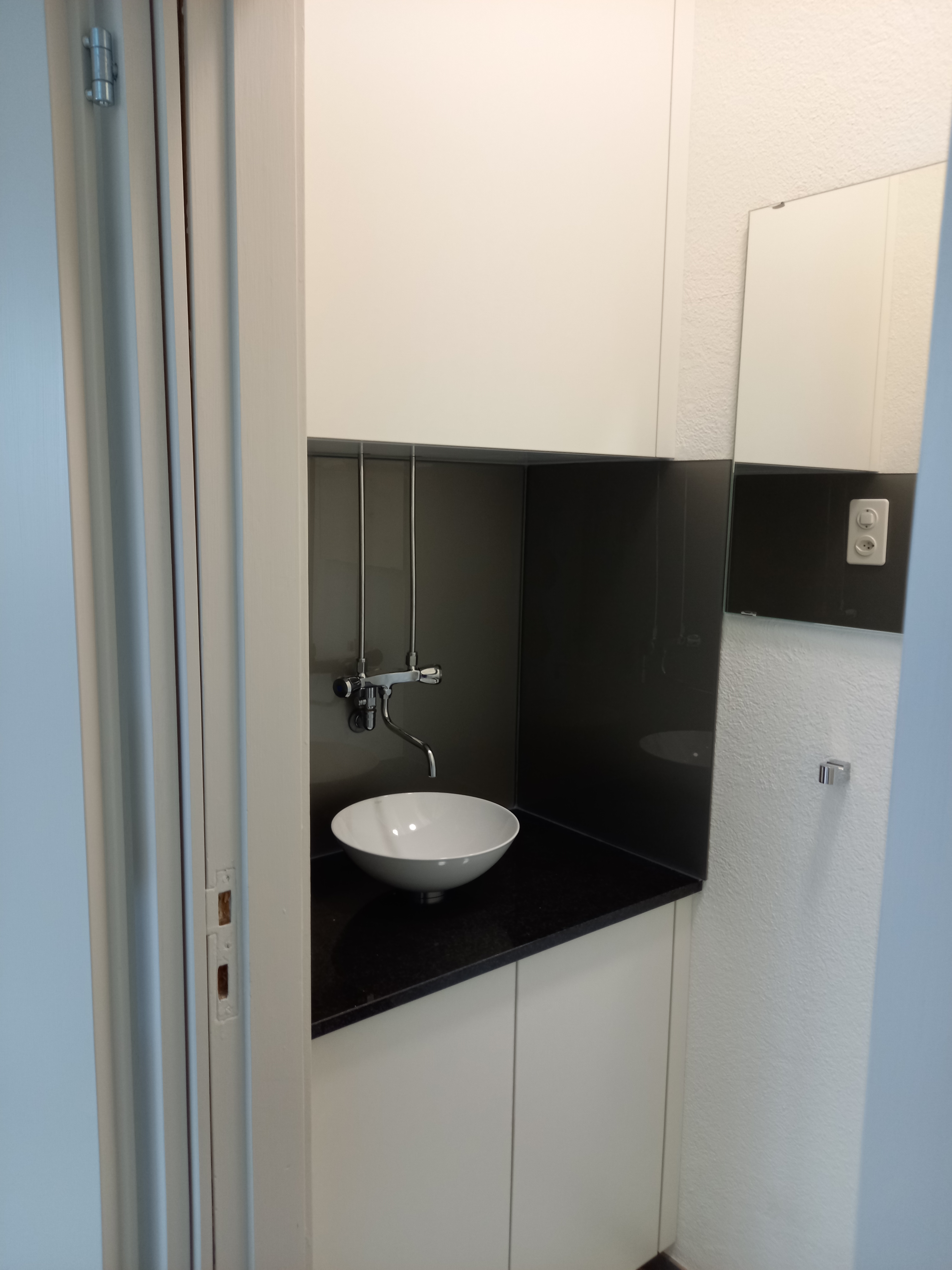 Modern small bathroom sink with round white basin, black countertop, and wall-mounted faucet inside white cabinetry.