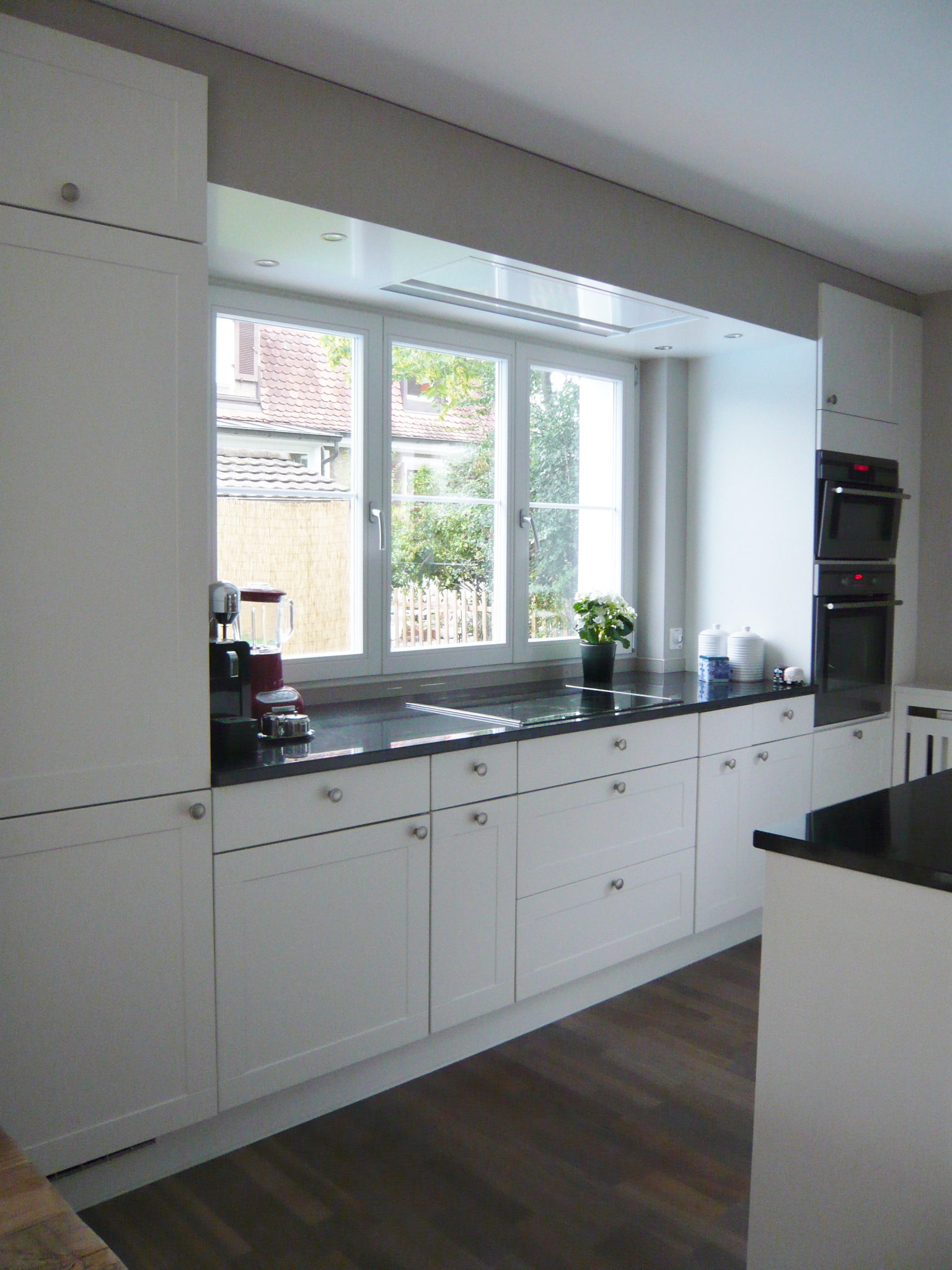 Modern kitchen with white cabinets, black countertops, built-in double oven, and a window overlooking greenery.