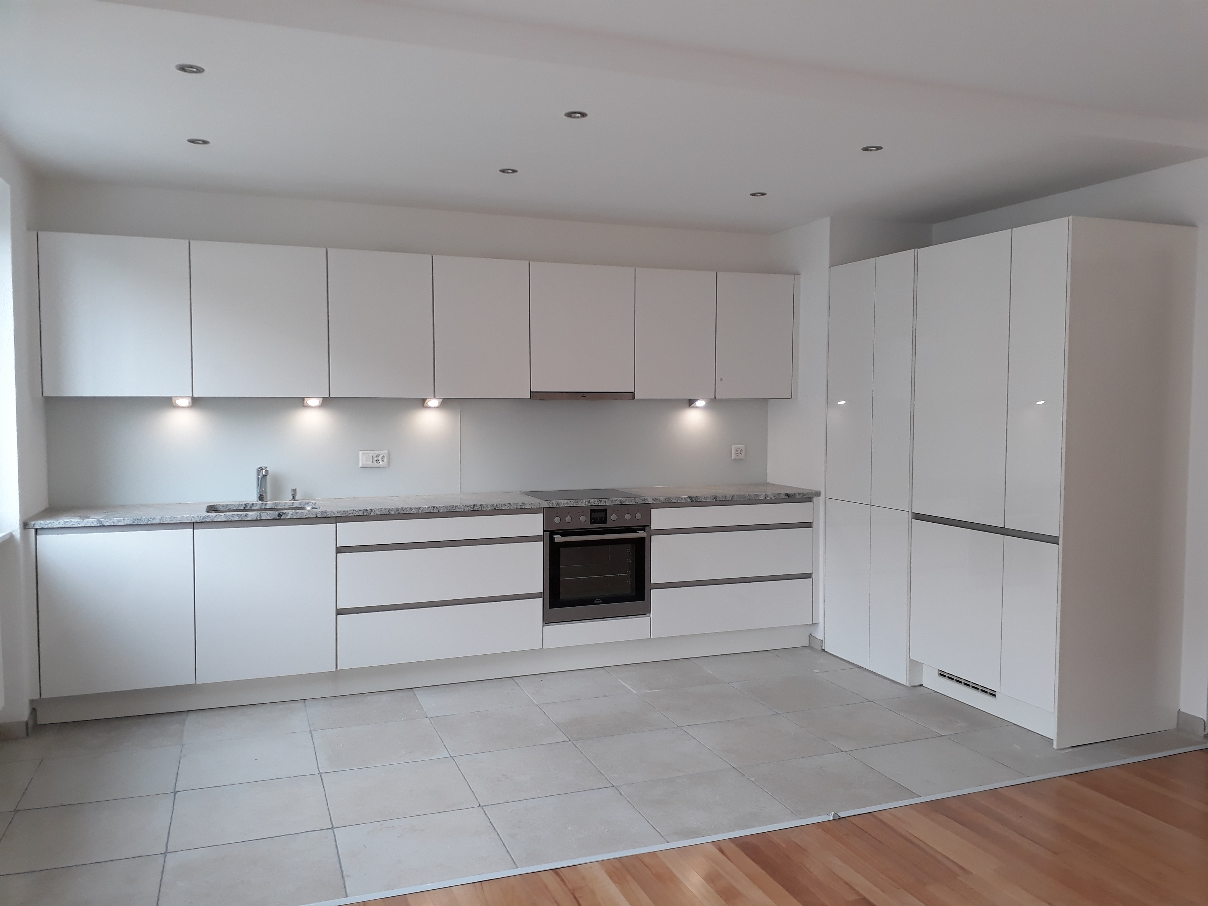 Minimalist modern white kitchen with marble countertop, built-in oven, stovetop, sink, and large storage cabinets.