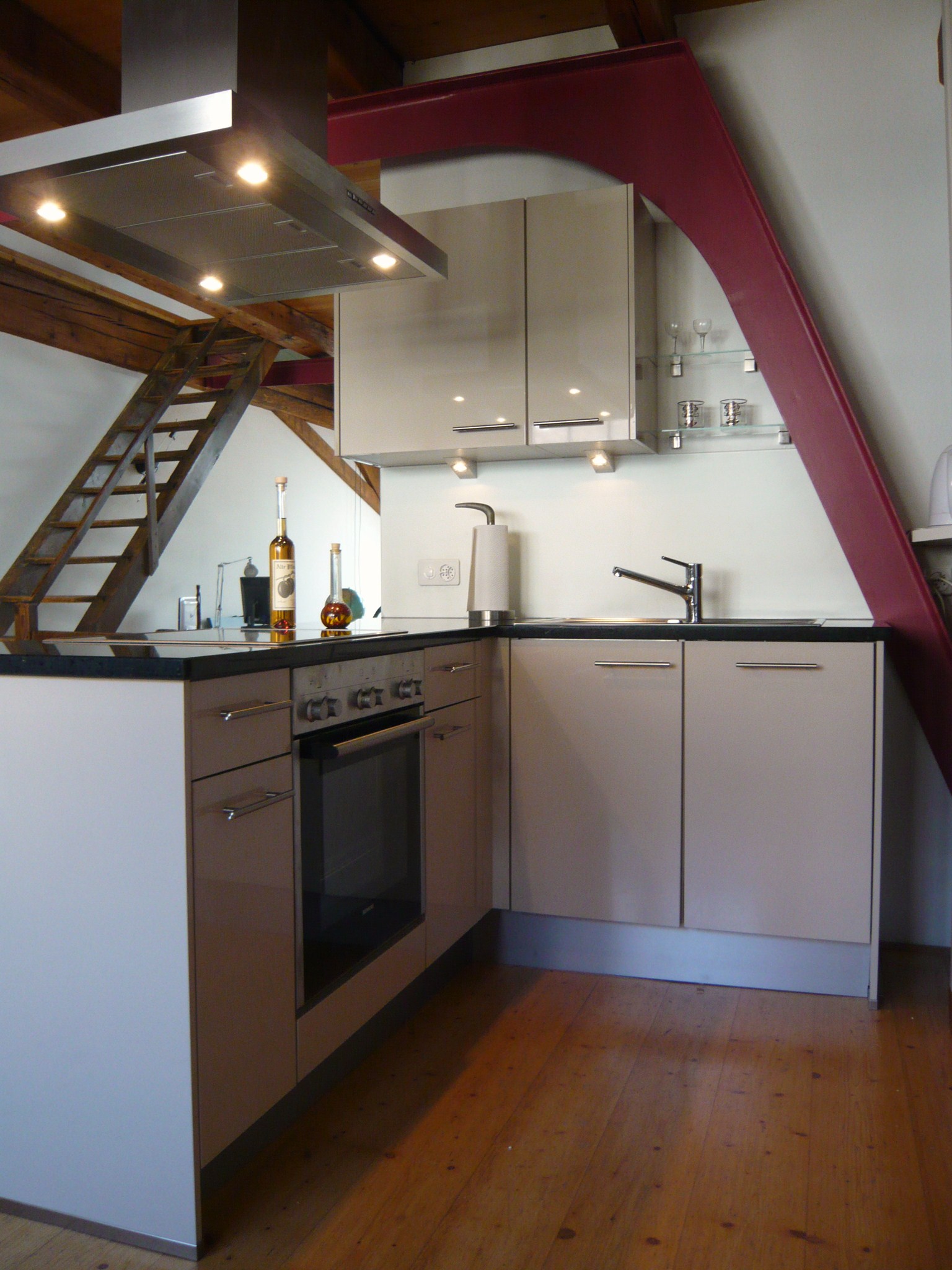 Modern kitchen corner with beige cabinets, black countertop, stovetop with hood, sink, and wooden floor under a slanted ceiling.