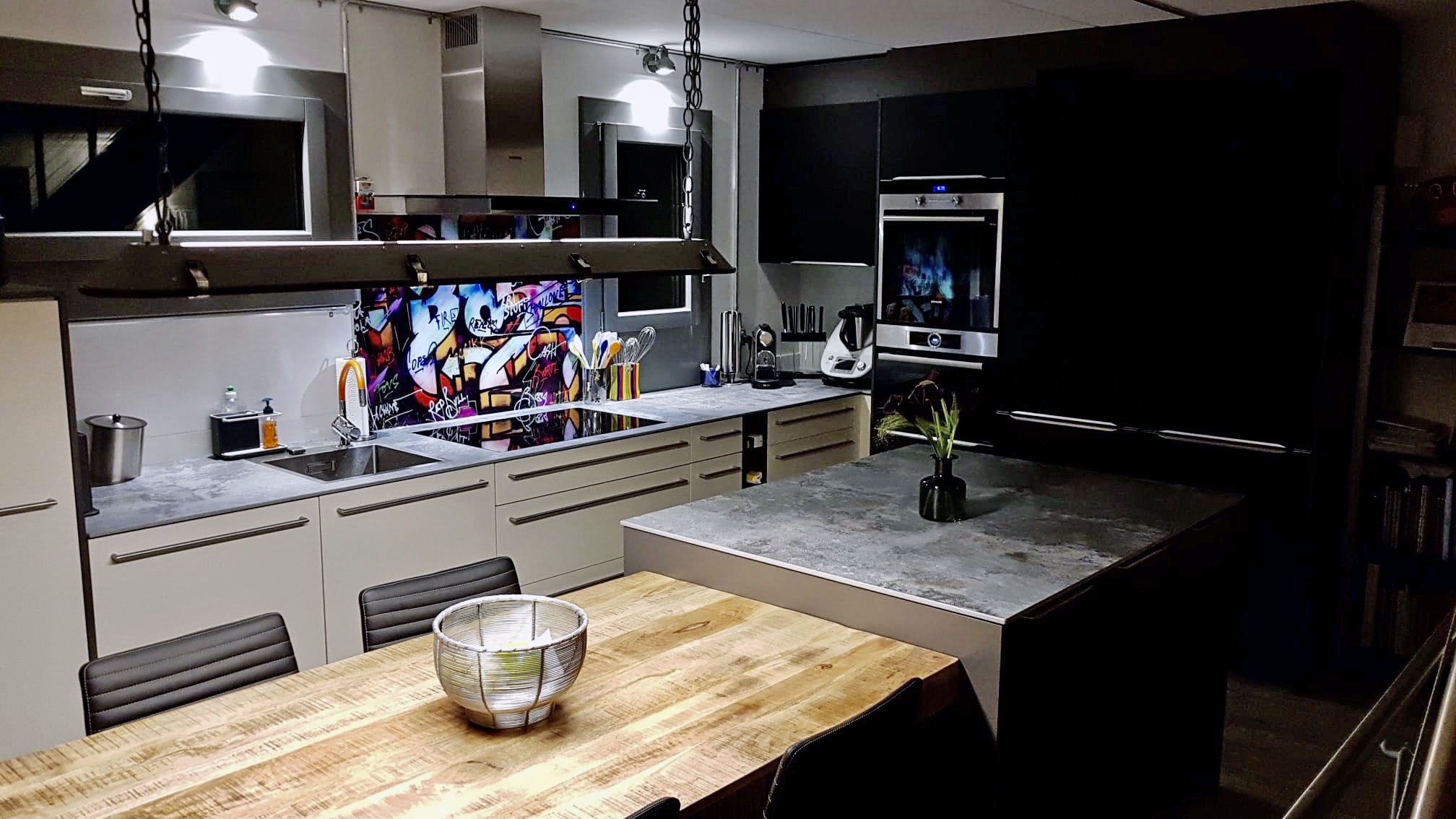 Modern kitchen with light-colored cabinets, colorful graffiti backsplash, wooden dining table, and dark island with a vase.