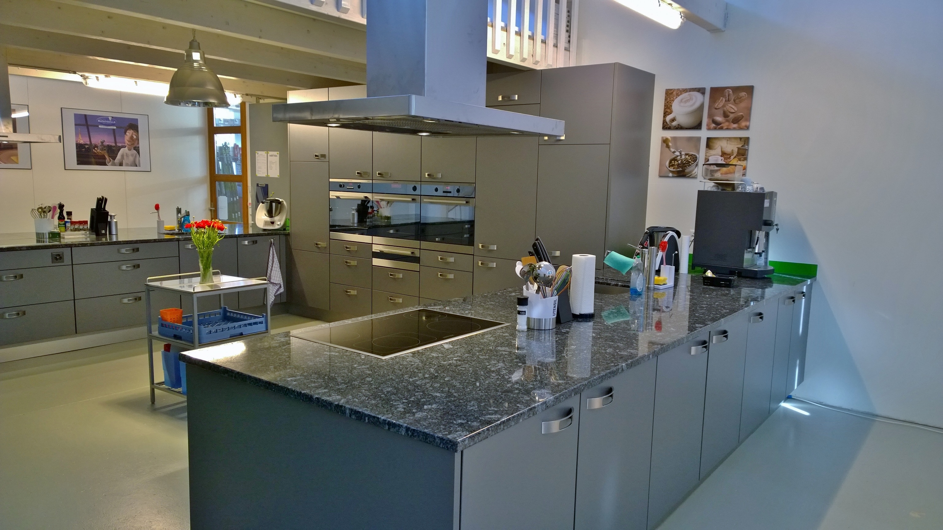 Modern kitchen with gray cabinets, a large granite island featuring an electric stove, utensils, and a coffee machine on the counter.
