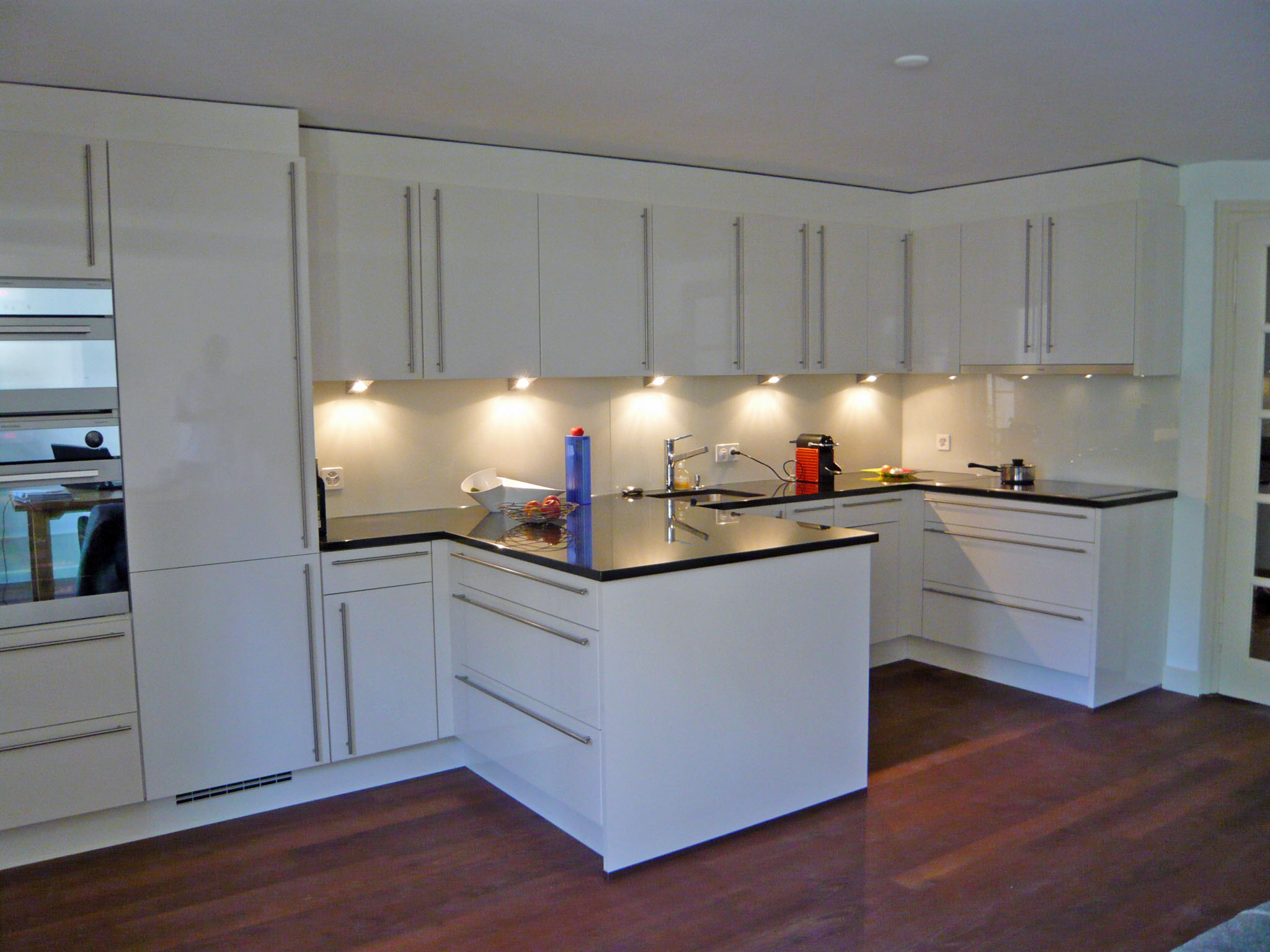 Modern kitchen with white cabinets, black countertops, under-cabinet lighting, and dark wooden floor.