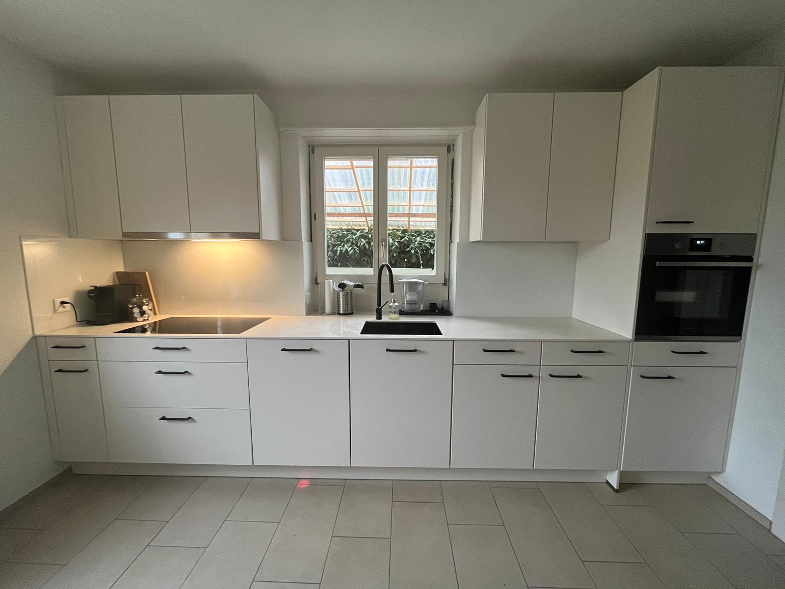 Modern white kitchen with black handles, built-in oven, electric stovetop, black faucet and sink under a window.