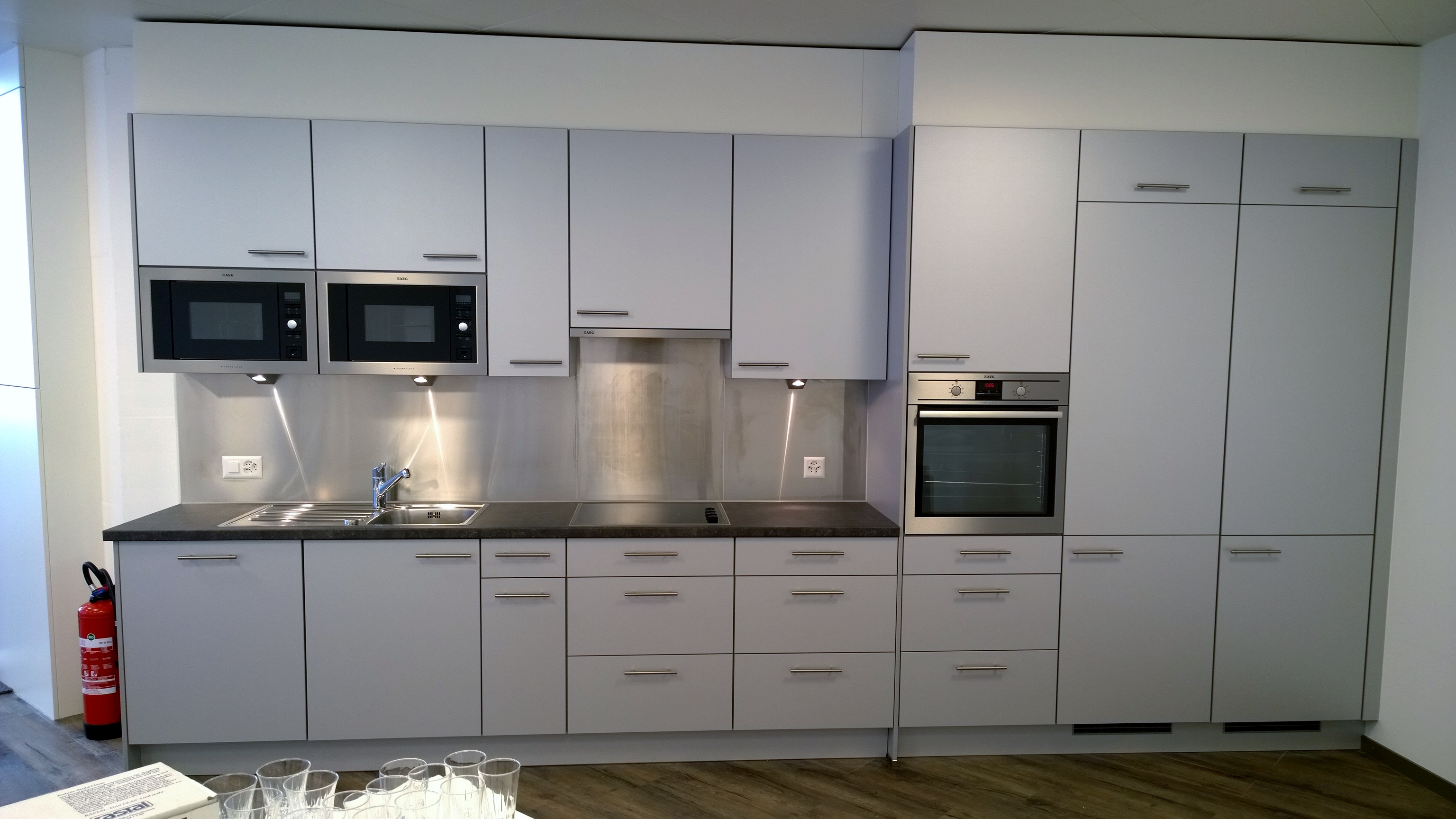 Modern kitchen with light gray cabinets, two built-in microwaves, stainless steel sink, stovetop, oven, and a fire extinguisher next to the counter.