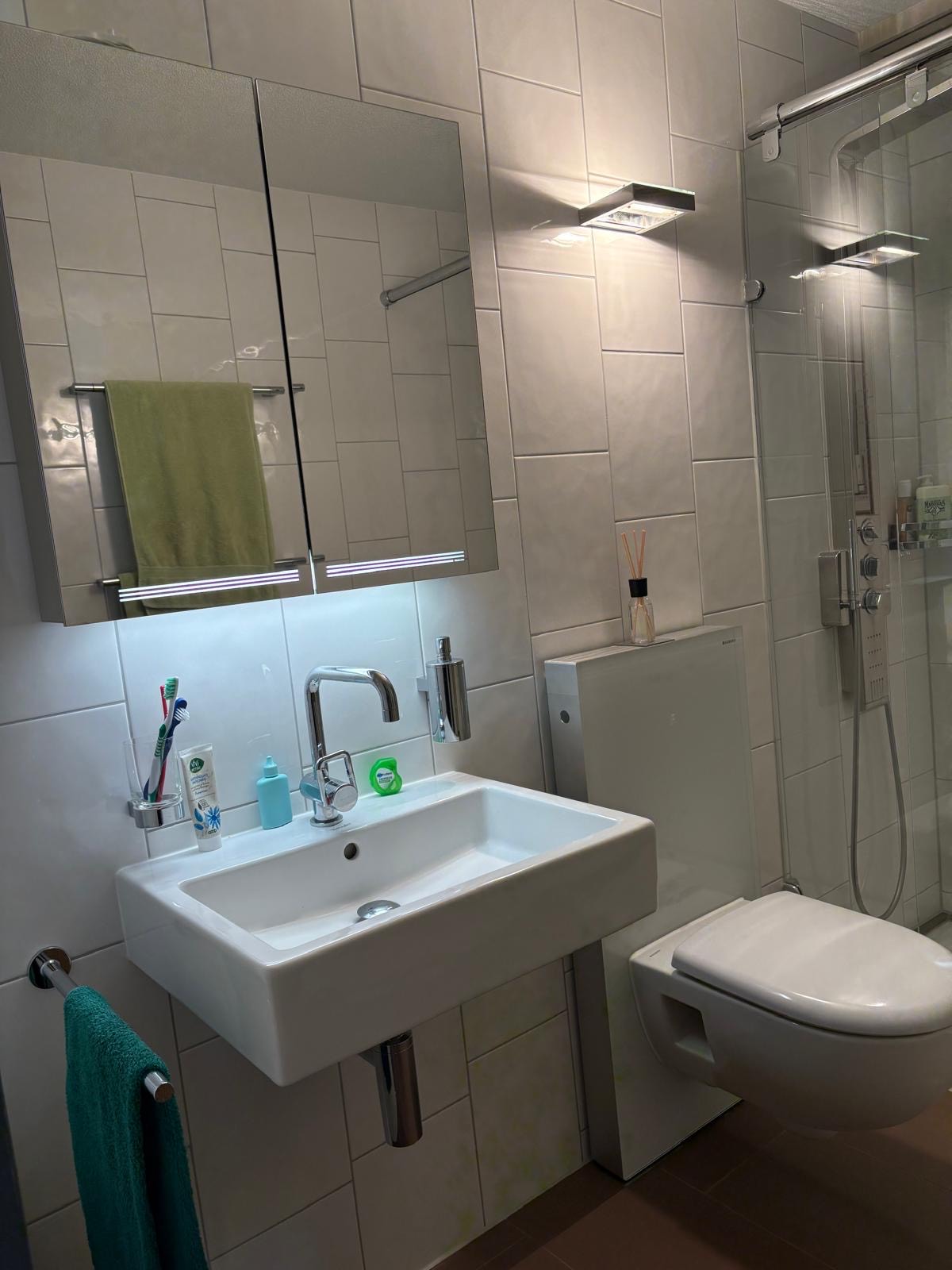 Modern bathroom featuring a white wall-mounted sink with toothbrushes and toiletries, a green towel hanging nearby, a wall-mounted toilet, and a glass shower enclosure.