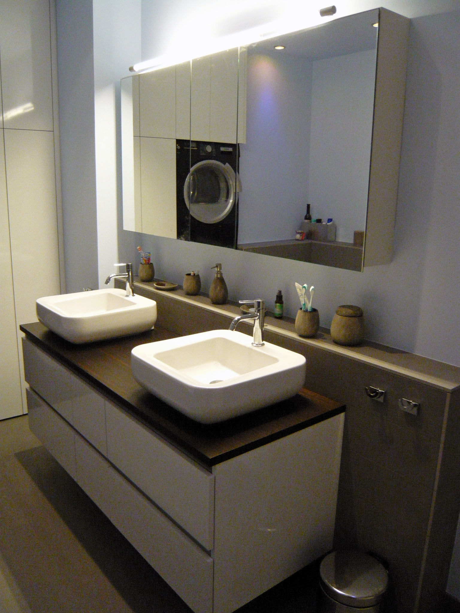 Modern bathroom with double square white sinks on a dark wood countertop, large mirrored cabinet, and various toiletry holders.