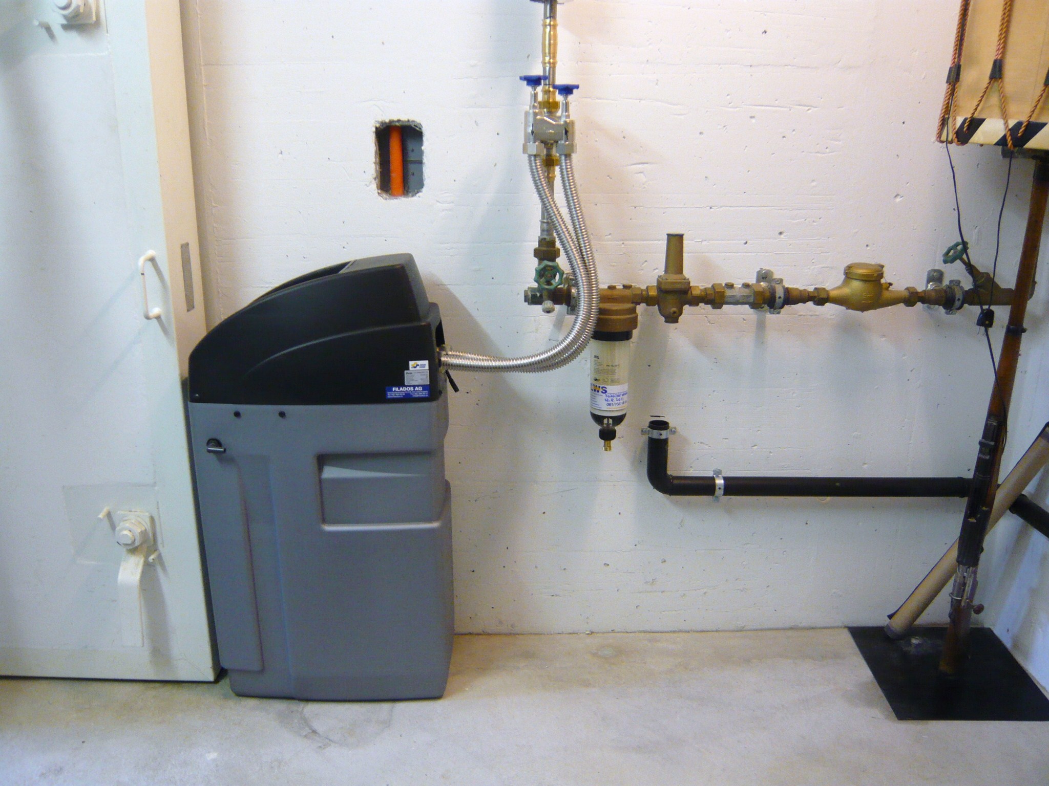 Gray and black water softener unit connected to plumbing pipes mounted on a white wall.