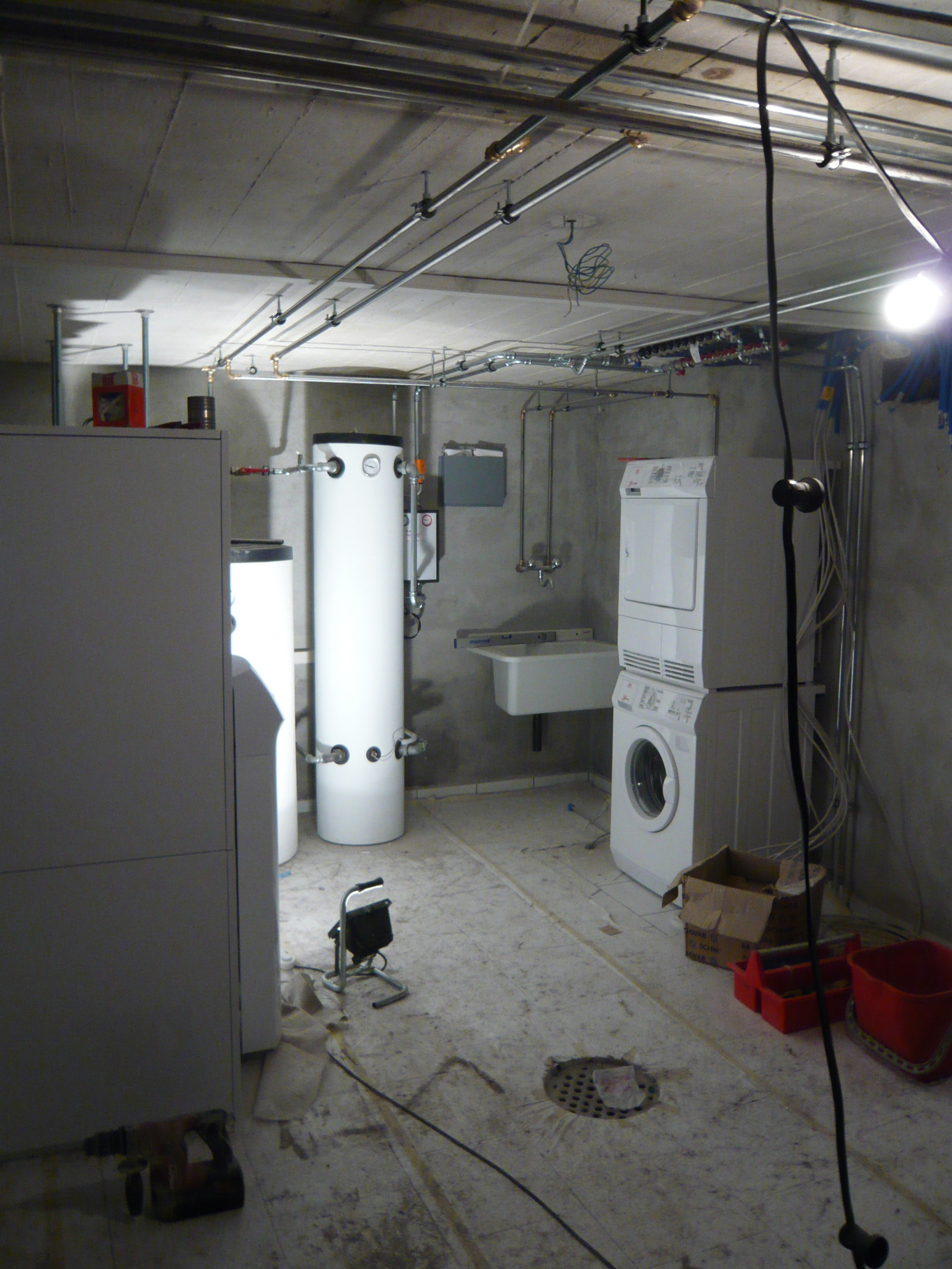 Basement laundry room with stacked washer and dryer, utility sink, water heater, exposed pipes, and construction tools on the floor.