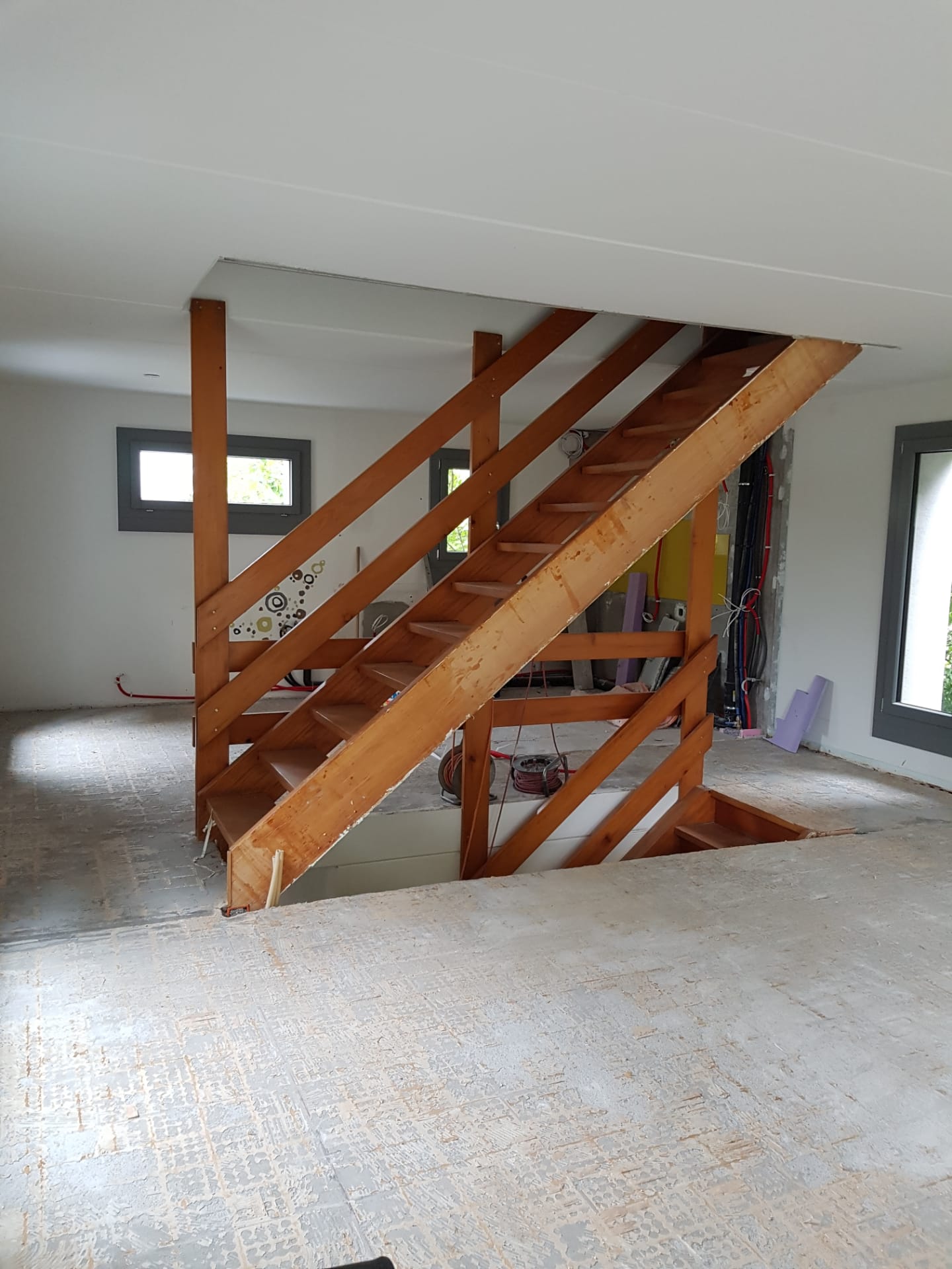 Interior under construction with unfinished floor and wooden staircase framed by vertical and diagonal supports.