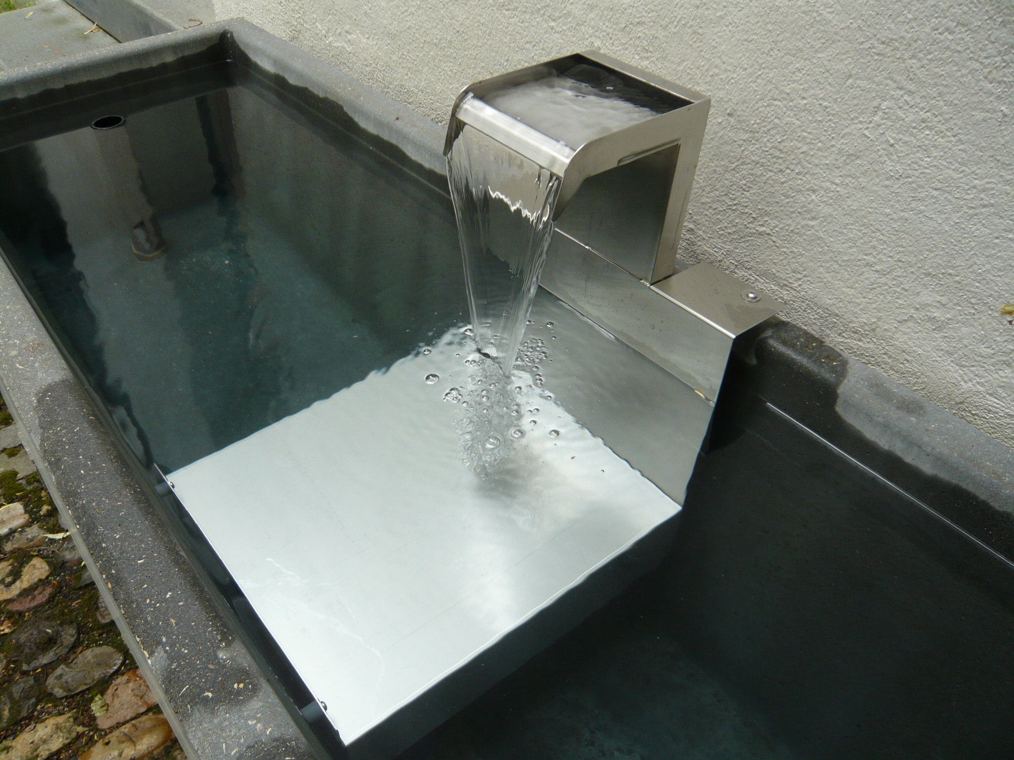Modern stainless steel water feature flowing into a rectangular stone basin filled with water.