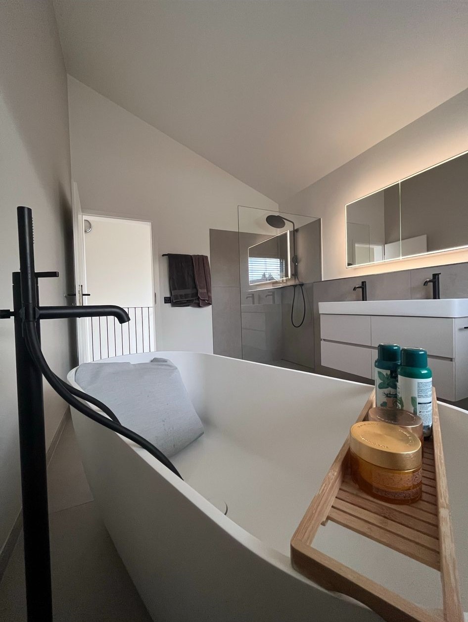 Modern bathroom with a white freestanding bathtub featuring a black floor-mounted faucet and a wooden bath tray holding toiletries, with a double sink vanity and mirror in the background.