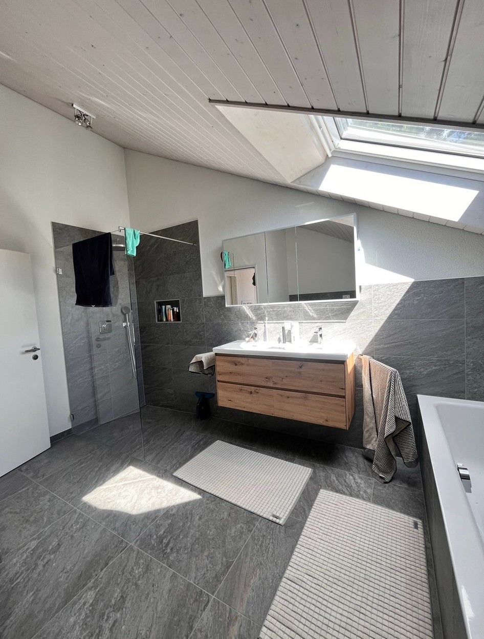 Modern bathroom with gray tiled floors and walls, wooden vanity with double sink, large mirror, walk-in glass shower, skylight, and two white bath mats.