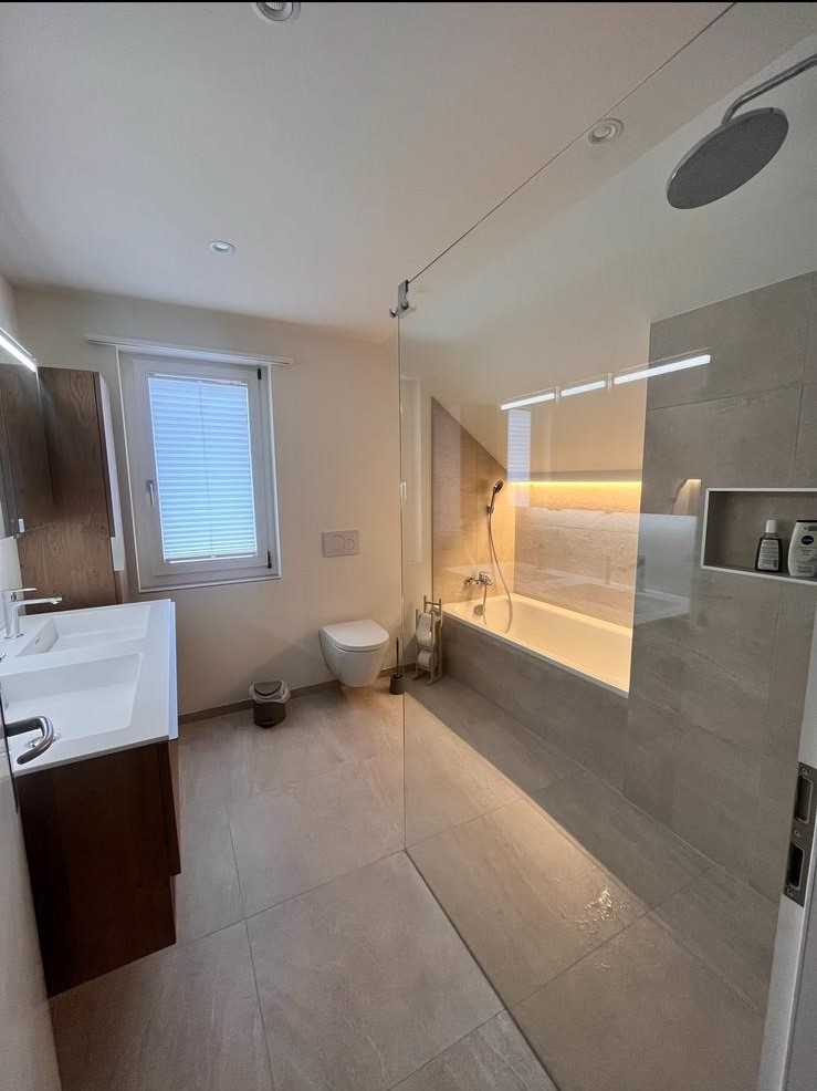 Modern bathroom with a double sink vanity, wall-mounted toilet, large window with blinds, glass-enclosed shower, and bathtub with warm underlighting.