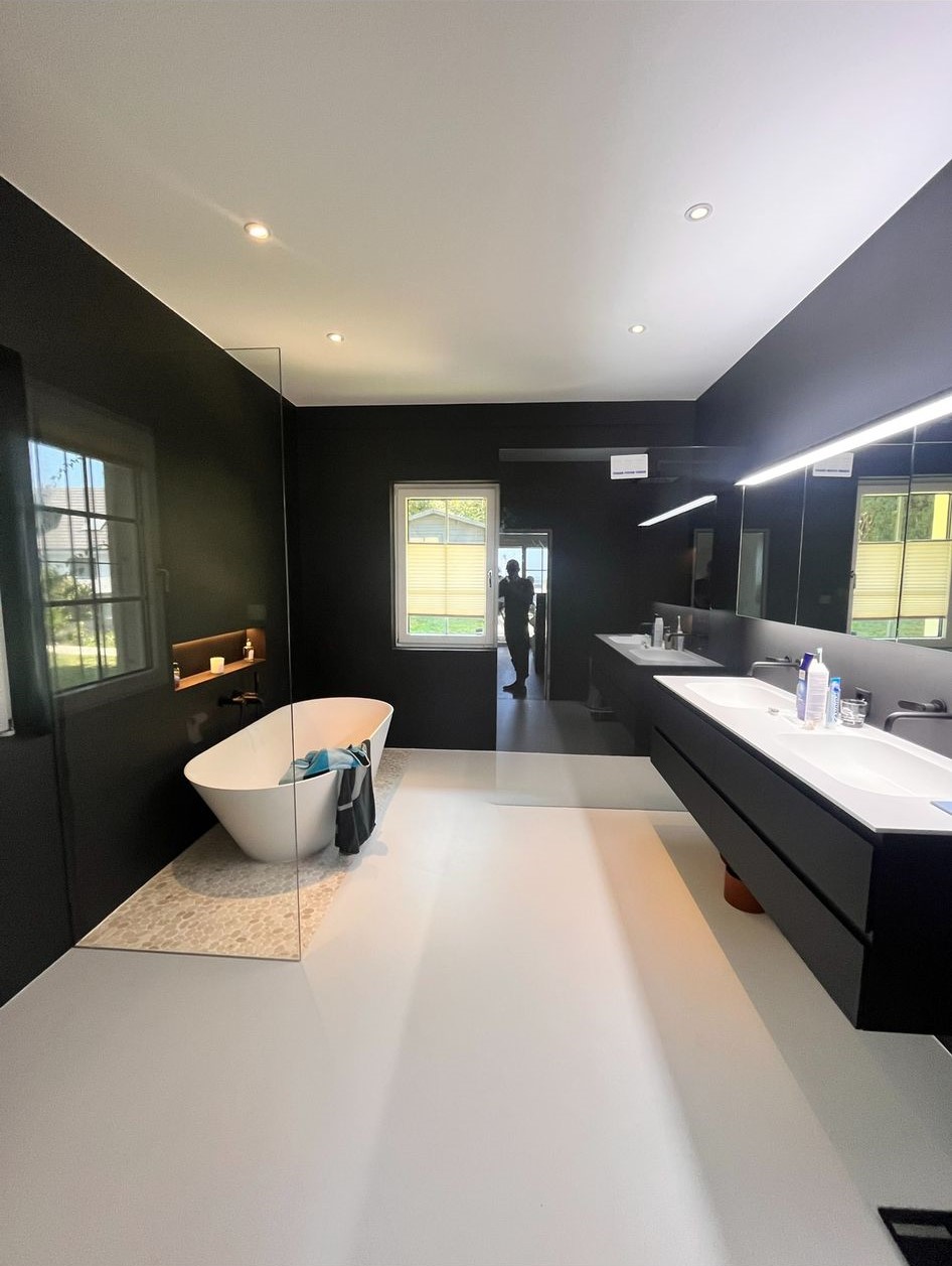 Modern bathroom with a white freestanding bathtub, black walls, dual sinks with a large mirror, and a window reflecting outdoor greenery.