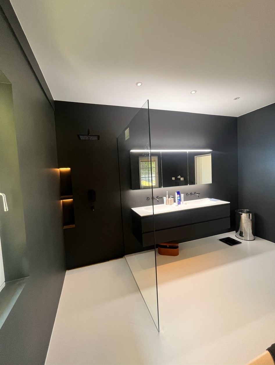 Modern bathroom with dark walls, a glass shower partition, wall-mounted double sink vanity, illuminated mirror, and a metallic trash bin.