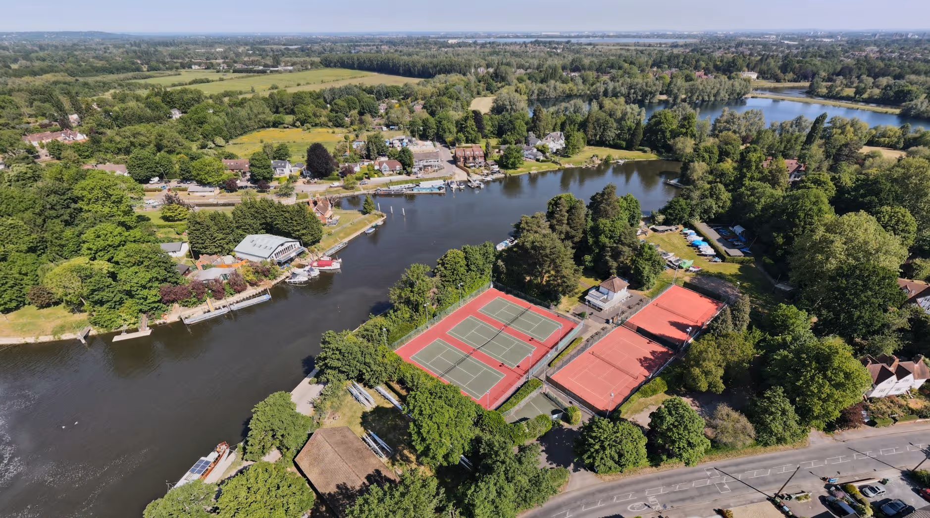 Aerial view of a river with surrounding greenery, residential houses, and tennis courts near the riverbank.