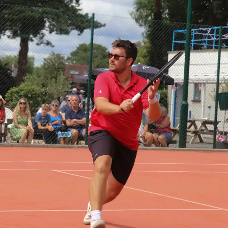 Man in red shirt and black shorts playing tennis on an outdoor clay court, preparing to hit a shot with a racket.