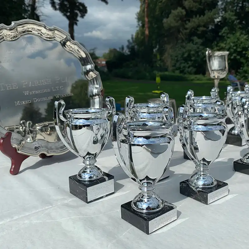 Several shiny silver trophies with black bases displayed on a white tablecloth outdoors with greenery in the background.