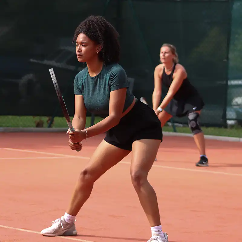 Two female tennis players on a clay court ready to receive a serve, one in the foreground holding a racket.