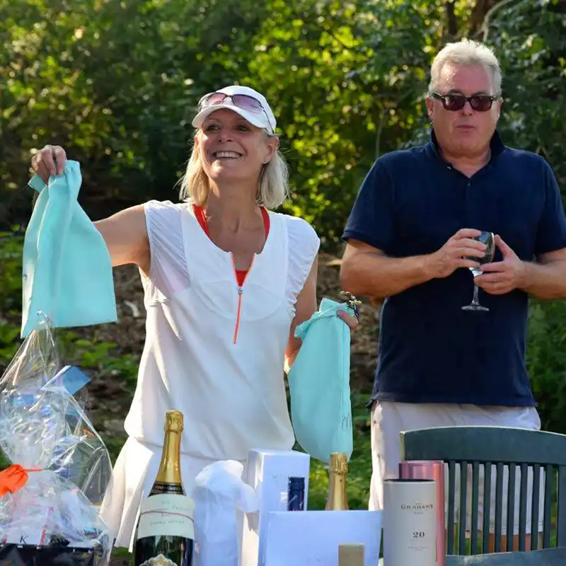 Smiling woman in white sportswear holding two light blue bags next to a man wearing sunglasses and holding a wine glass outdoors.