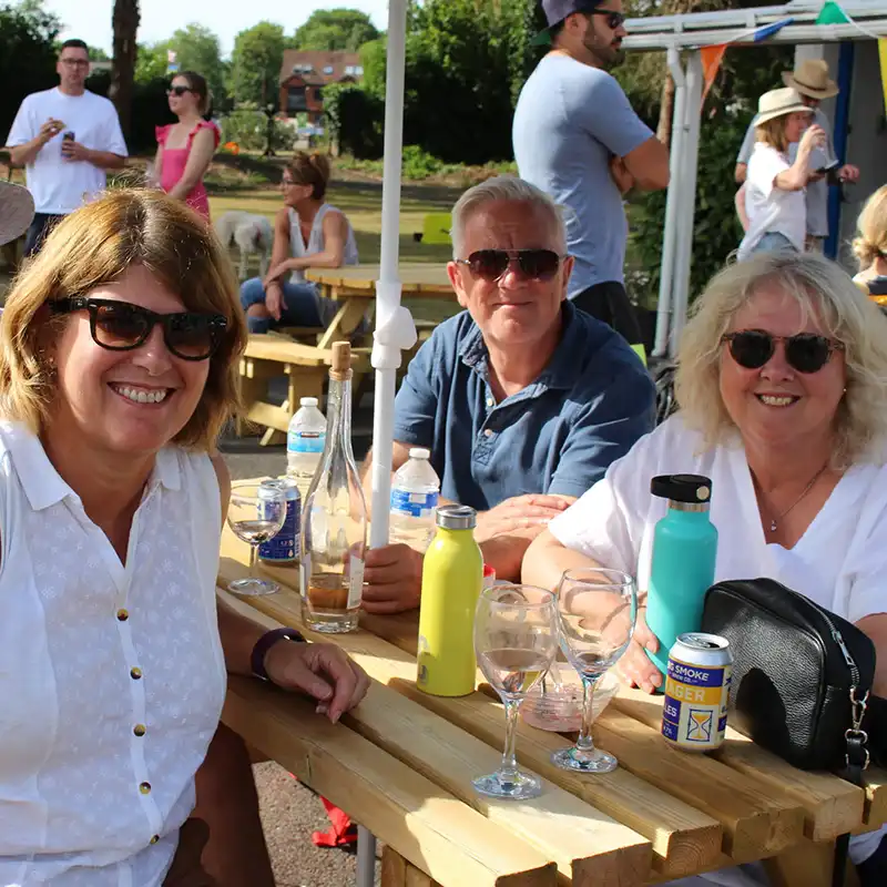 Three adults wearing sunglasses sitting and smiling around a wooden picnic table with drinks during an outdoor gathering.